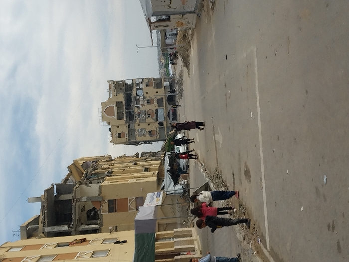 This image depicts a street scene in Khan Yunis, Palestinian Territory, under a partly cloudy sky. The scene is dominated by multi-story residential or commercial buildings exhibiting significant structural damage, with collapsed sections, exposed interiors, and missing facades.
On the left, a light-colored building with multiple windows shows severe damage to its upper floors, including exposed rebar and broken concrete. Below it, a makeshift stall with a green and white awning is visible on the sidewalk. Further down the street on the right, another multi-story building, also light-colored and featuring an arched architectural detail near its roofline, displays extensive damage with numerous gaping holes where windows or walls once were.
The asphalt road in the foreground is strewn with debris and dirt, with faded white lane markings, including an arrow pointing forward. On the left sidewalk, a group of approximately four children are standing or interacting, dressed in everyday clothing; one child wears a dark jacket, another a pink top, and two others are visible. Further down the street, an adult in dark clothing walks away from the viewer on the right side of the road, and other individuals and vehicles are partially visible in the distance, suggesting ongoing activity despite the evident destruction. Debris piles and smaller, temporary structures are scattered along the sides of the road.