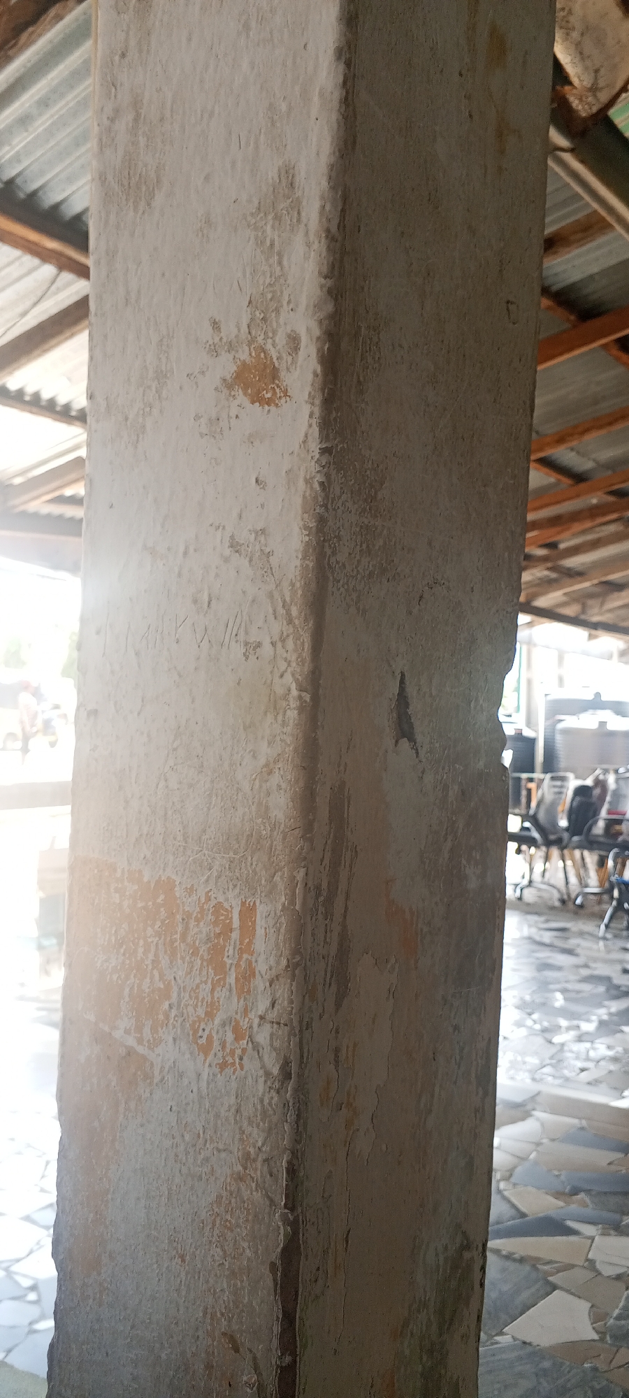 The image shows a close-up of a textured concrete pillar. The pillar has patches of peeling paint and some faint markings or carvings on its surface. The background is blurred, but suggests an outdoor or semi-outdoor setting with a corrugated metal roof structure visible above. Sunlight is streaming in from the left, illuminating part of the pillar and casting shadows. In the further background, there are glimpses of people and chairs, indicating a public or communal space. The floor appears to be tiled with irregularly shaped pieces. The scene is likely located in Jalingo, Nigeria, given the context provided.