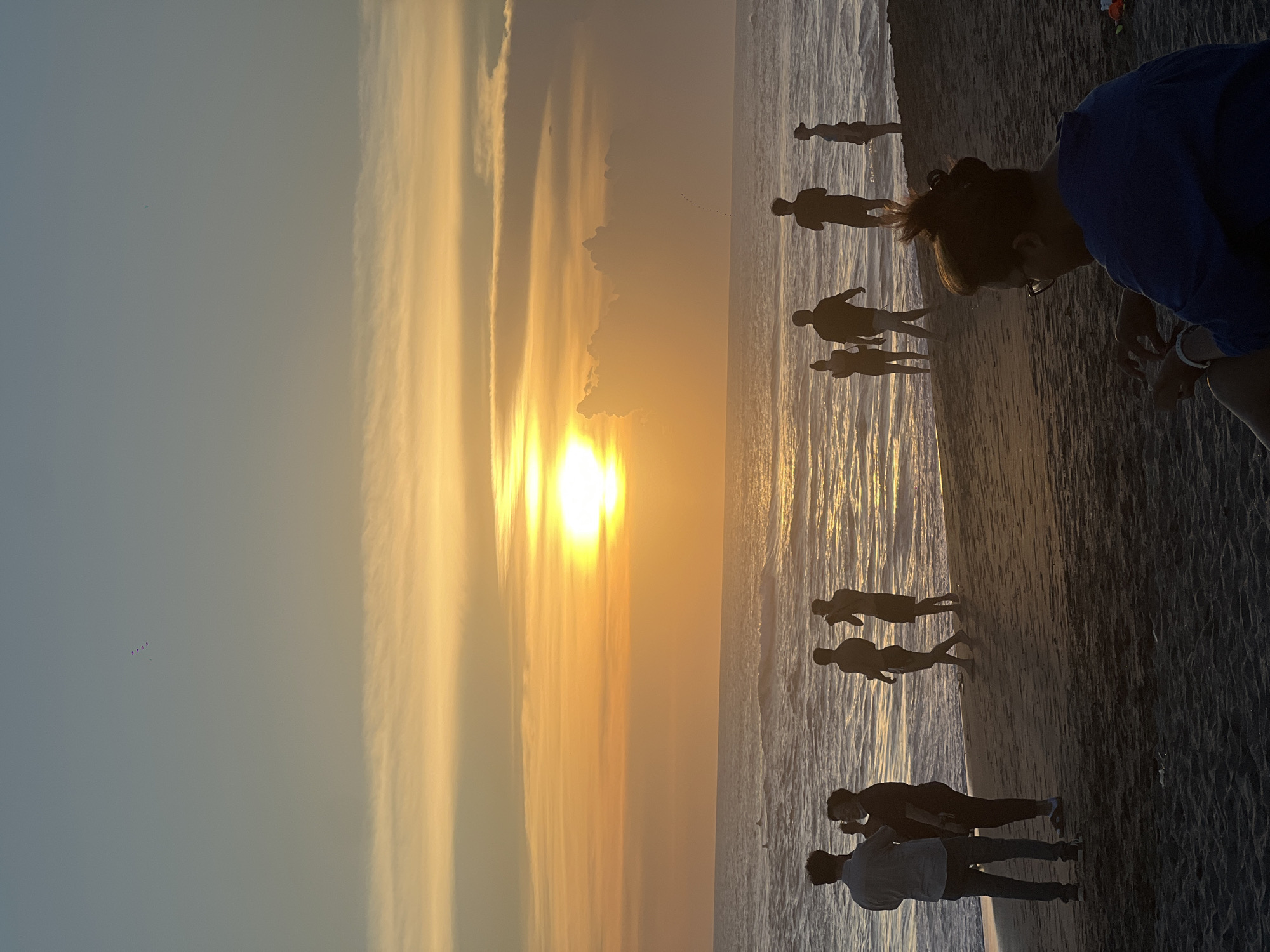 The image shows a sunset scene on a beach in North Kuta, Indonesia. Several people are on the beach, silhouetted against the bright sun and the ocean. One person is closer to the camera, facing down towards the sand. The sky is a mix of blue and orange colors, with sunlight reflecting on the water.