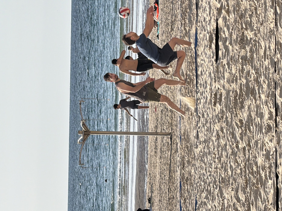 The image captures a sunny beach scene in Tel-Aviv, Israel, with the Mediterranean Sea in the background. The foreground consists of light-colored sand, marked by a faint blue line indicating a court boundary. Five males are visible. In the mid-ground, two adult males are actively engaged in a beach volleyball game. On the right, a male in a dark blue t-shirt and dark shorts is seen lunging forward, arms extended, making contact or preparing to contact a red, white, and blue volleyball suspended in the air. His right foot is planted, causing sand to spray upwards. To his left, another male in a grey sleeveless shirt and olive-green shorts stands, observing the play, with sand disturbed around his left foot. Behind these players, a shirtless male in dark shorts is partially visible, with his right hand near his face. Further back, closer to the water's edge, a younger male in a dark t-shirt and shorts holds a paddle or small racket. A metallic pole structure with outward-curving arms and hanging lines stands to the left of the central activity. The sea exhibits light waves under a clear, pale blue sky.