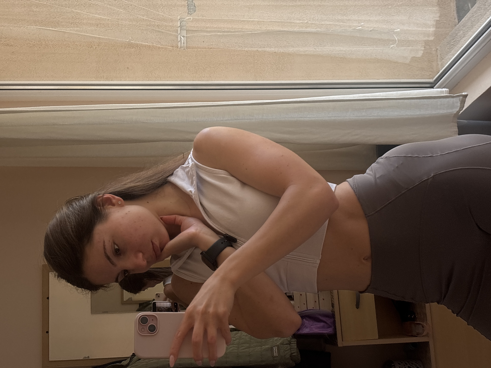 A young woman with brown hair pulled back, wearing a white cropped athletic top and gray leggings, takes a selfie in a room with a mirror and a window. She is holding a pink smartphone in her left hand and her right hand is resting on her chin. A dark-colored smartwatch is visible on her left wrist. Behind her, a wardrobe with clothes and a chest of drawers are visible. The window is covered by light-colored curtains. The lighting suggests it is daytime. The location in Varna, Bulgaria is not discernible from the image.