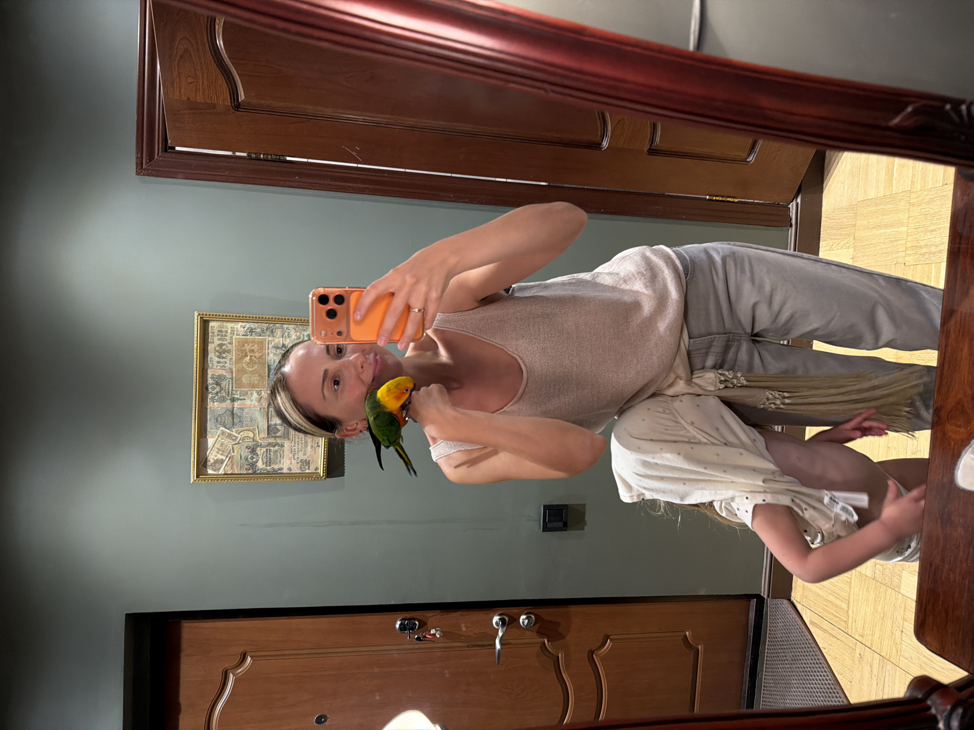 A female individual with light brown hair, dressed in a light-colored tank top and gray sweatpants, takes a mirror selfie. She holds an orange-cased smartphone in her right hand, capturing her reflection and the colorful parrot perched on her left shoulder, with the parrot's head near her neck. A small child, wearing a light-colored outfit, is visible to the right of the woman, partially obscured by the woman's body and a wooden surface.
The setting appears to be an indoor space with a teal-colored wall. On the wall, behind the woman, hangs a framed picture with what looks like old currency or stamps. To the right, a wooden door with intricate carvings is visible, and below it, part of a wooden floor with a parquet pattern can be seen. Another wooden door is reflected in the lower part of the mirror. A dark rectangular object, possibly a light switch or outlet, is on the wall behind the woman's mid-section. The reflection in the mirror also shows parts of what appears to be a dark wooden frame or furniture. There is no visible text.