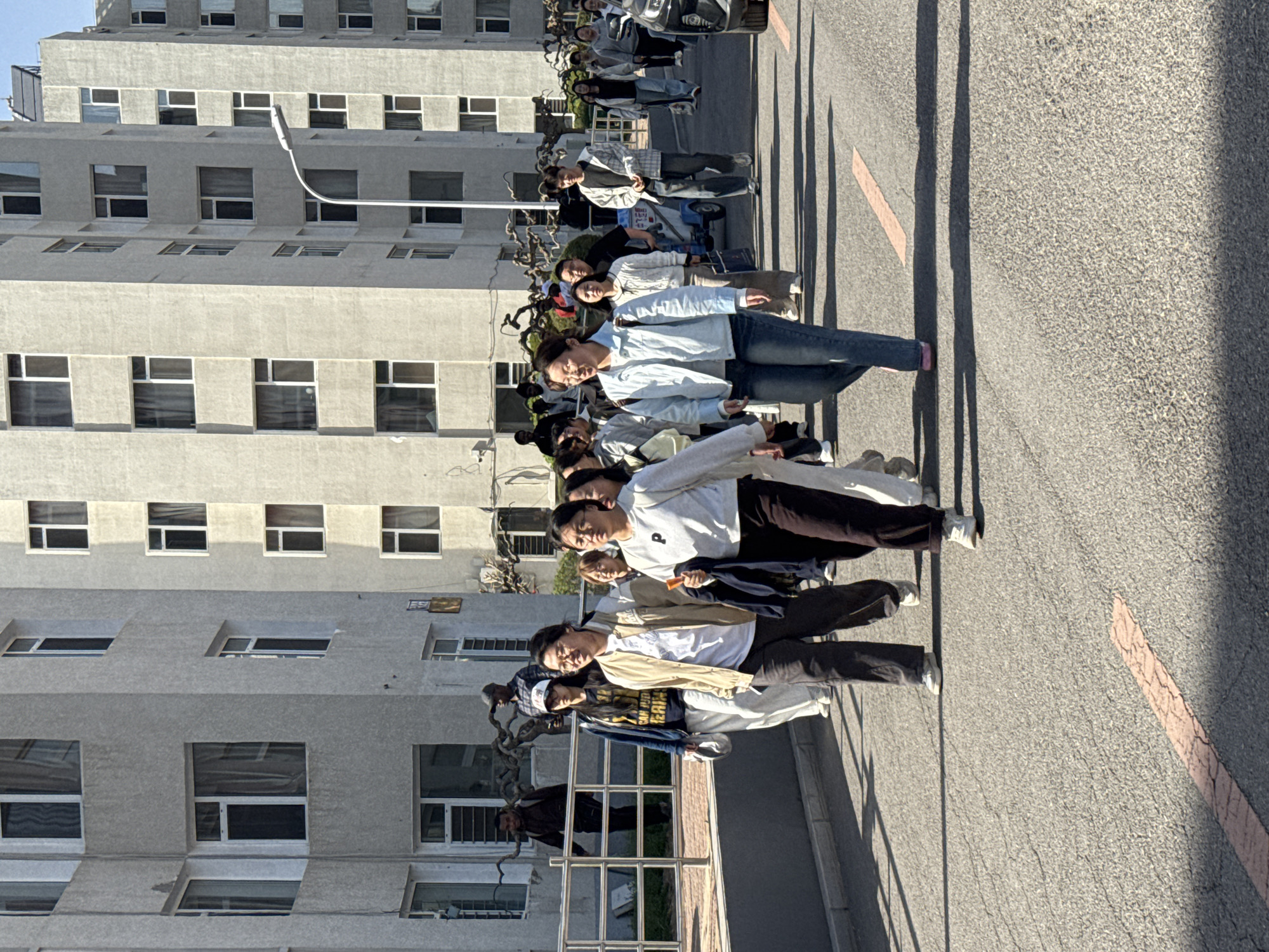 The image captures a group of people, primarily young adults, standing outdoors in what appears to be a line or casual gathering. They are positioned on a paved area, possibly a road or pathway, with a tall, multi-story building to their left and an open expanse of asphalt to their right. Most individuals are dressed in casual attire, including jeans, t-shirts, and light jackets, and some are looking towards the camera. Their expressions seem relaxed.
Long shadows cast by the people and other objects stretch across the ground towards the right, indicating either early morning or late afternoon with bright, direct sunlight. The weather appears clear and sunny.
The background features a large, grey concrete building with numerous rectangular windows. Adjacent to the building, a street light pole extends into the scene. Several bicycles or scooters are parked near the building, behind the group of people. The paved ground shows some cracks, and a faded, cracked red line is visible on the asphalt to the far right.
On a white sign attached to a parked scooter or stand near the people, the numbers "11-2" and "43" are visible, along with some indistinct Chinese characters. The overall setting suggests an urban or campus environment in Zhongtun, China.