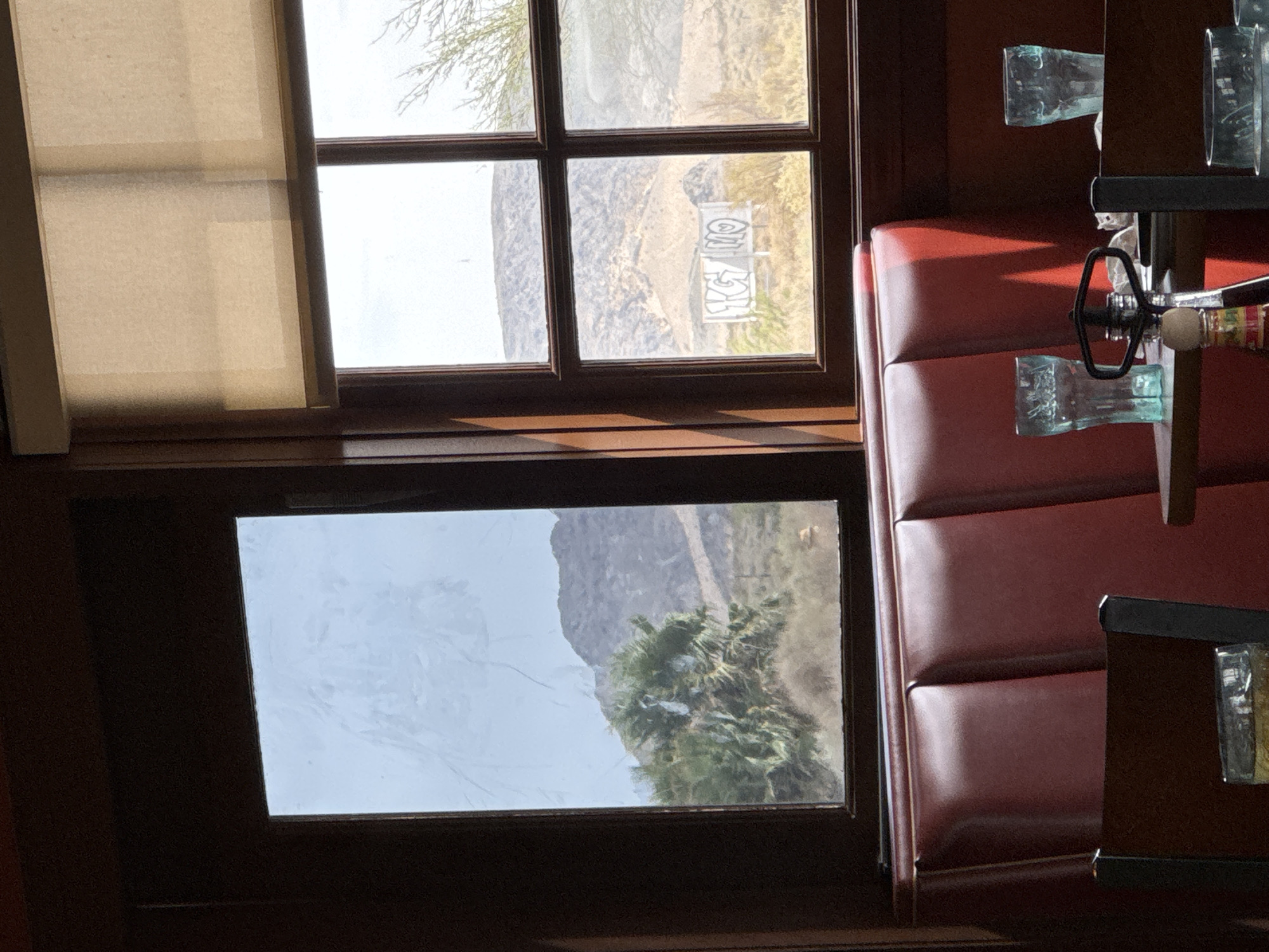 The image shows a view from inside a building, likely a diner or restaurant, looking out through two windows. The interior features a red leather booth and wooden framing. Outside, the landscape is arid, with mountains in the background and sparse vegetation, including a large, leafy tree on the left. In the right window, a white structure with graffiti on it is visible. The sky is overcast. The scene is located in Baker, California, United States of America.