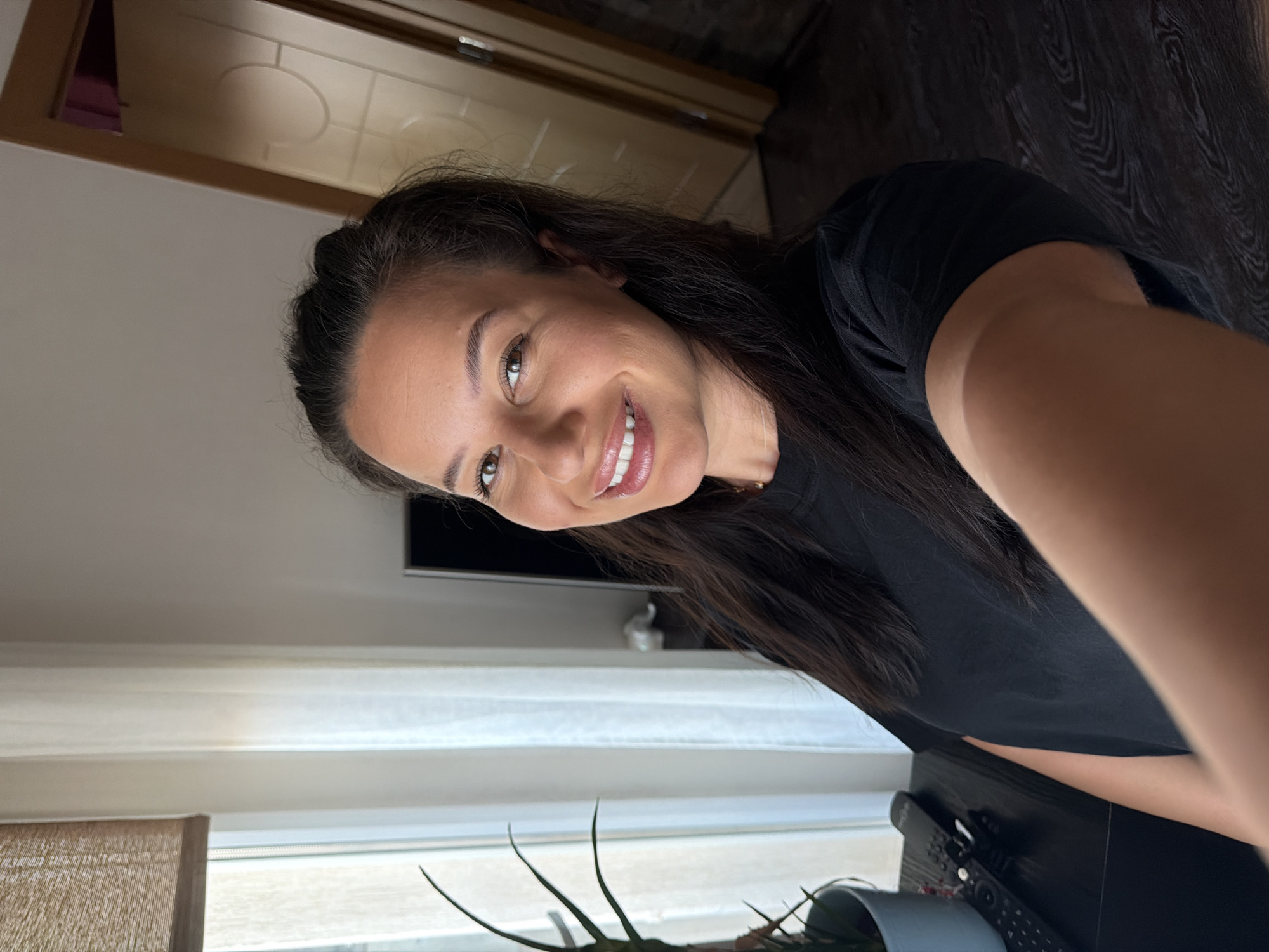 The media file shows a close-up, selfie-style shot of a woman with long, dark hair, wearing a black t-shirt. She is smiling and looking towards the camera. Her skin tone is light to medium, and her eyes are brown. She appears to be indoors, with a window with blinds visible in the background. The lighting suggests it could be daytime. Behind her, a wooden floor and a doorway with some decorative paneling can be seen. On a surface in the lower right corner, a remote control is visible. The overall mood is casual and friendly, conveyed by her smile. The image is oriented sideways.