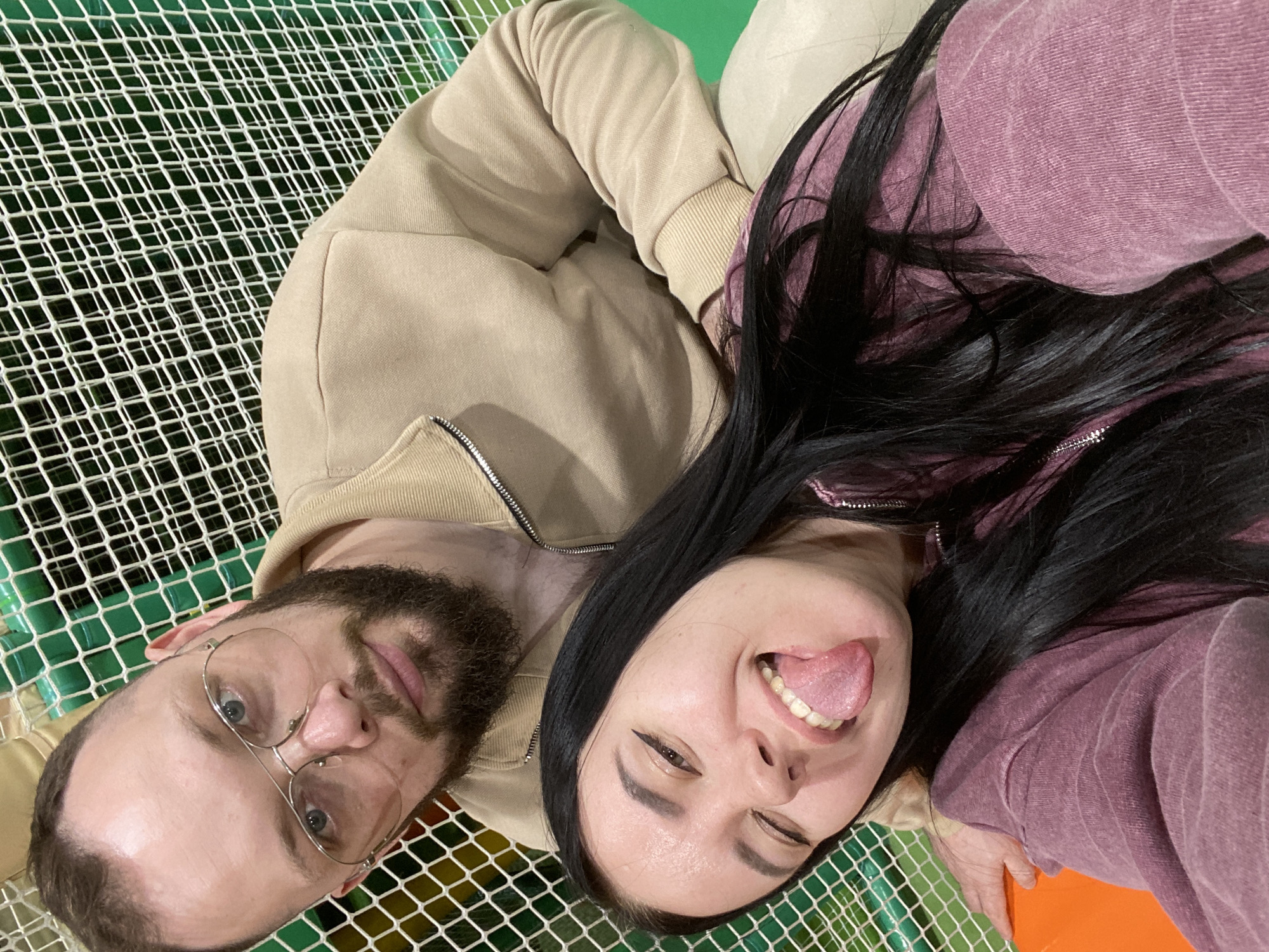 The image shows two people lying down, likely inside an indoor play area or amusement center, with a white net visible in the background, suggesting a play structure. A man with a beard and glasses is on the left, and a woman with long black hair is on the right. The woman is sticking her tongue out. The location is purportedly Perm, Russia, but there are no identifiable landmarks visible to confirm. Both are wearing casual attire, jackets or sweatshirts, and are likely taking a selfie or having their picture taken. The play area has padding or soft flooring visible in green, orange, and yellow.