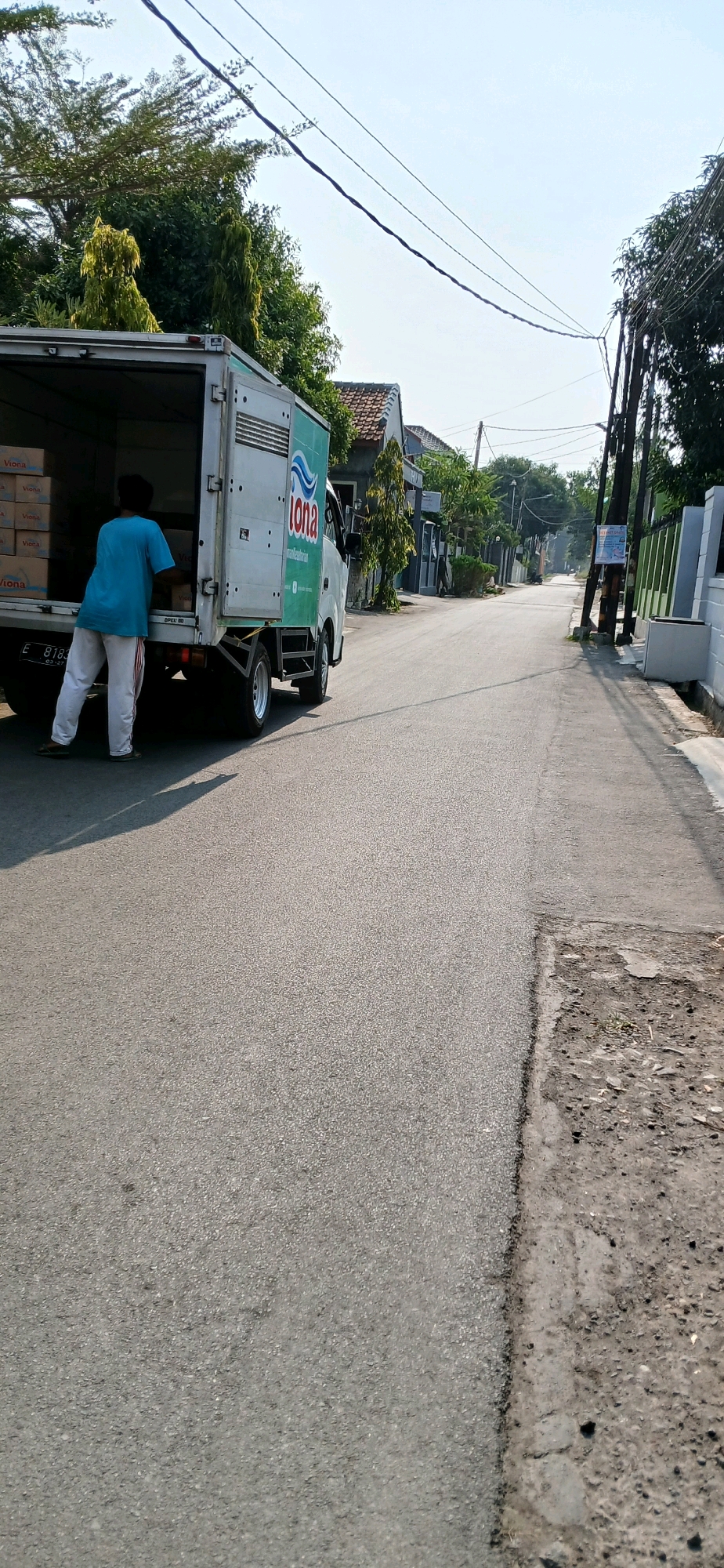 A person wearing a blue t-shirt and white pants is standing next to the open back of a white delivery truck in Sutawinangun, Indonesia. The truck is partially loaded with boxes, some of which are labeled "Viona". The truck is parked on the side of a paved road lined with houses and trees. The sky is clear and bright.