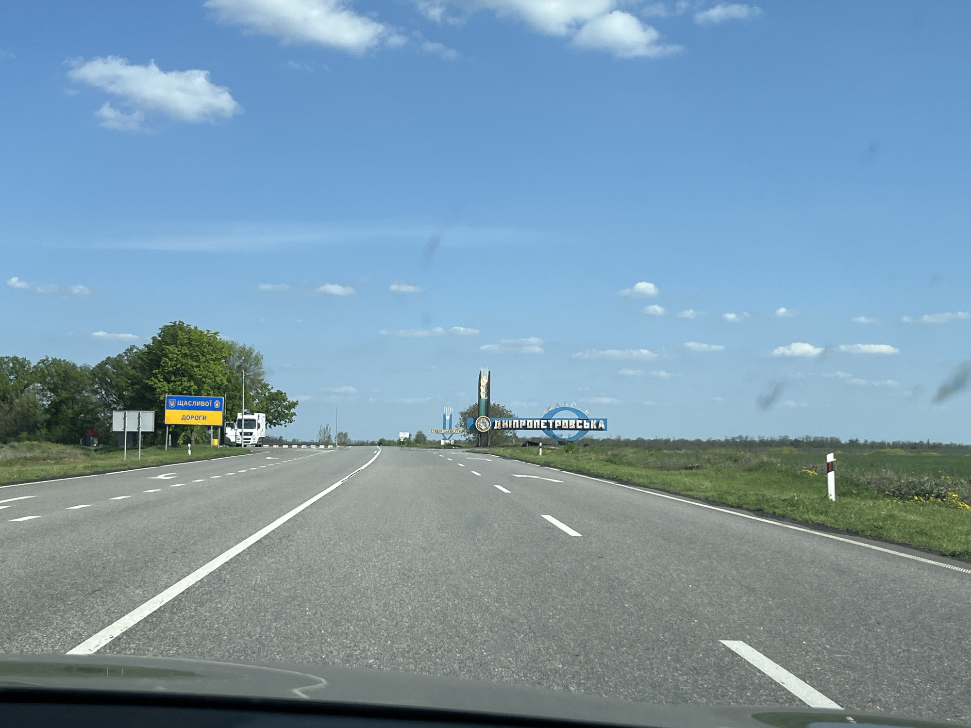 The image shows a straight, paved highway in a rural area. The road leads towards a sign indicating "Dnipropetrovska" in Ukrainian, suggesting the location is near the Dnipropetrovsk region. Another sign on the left side of the road reads "SHASLIVOI DOROGI" in Ukrainian. A white truck is parked near this sign. The sky is blue with scattered clouds. Vegetation borders both sides of the road. The image appears to be taken from inside a vehicle. The location is near Hurivka, Ukraine, based on the signs.