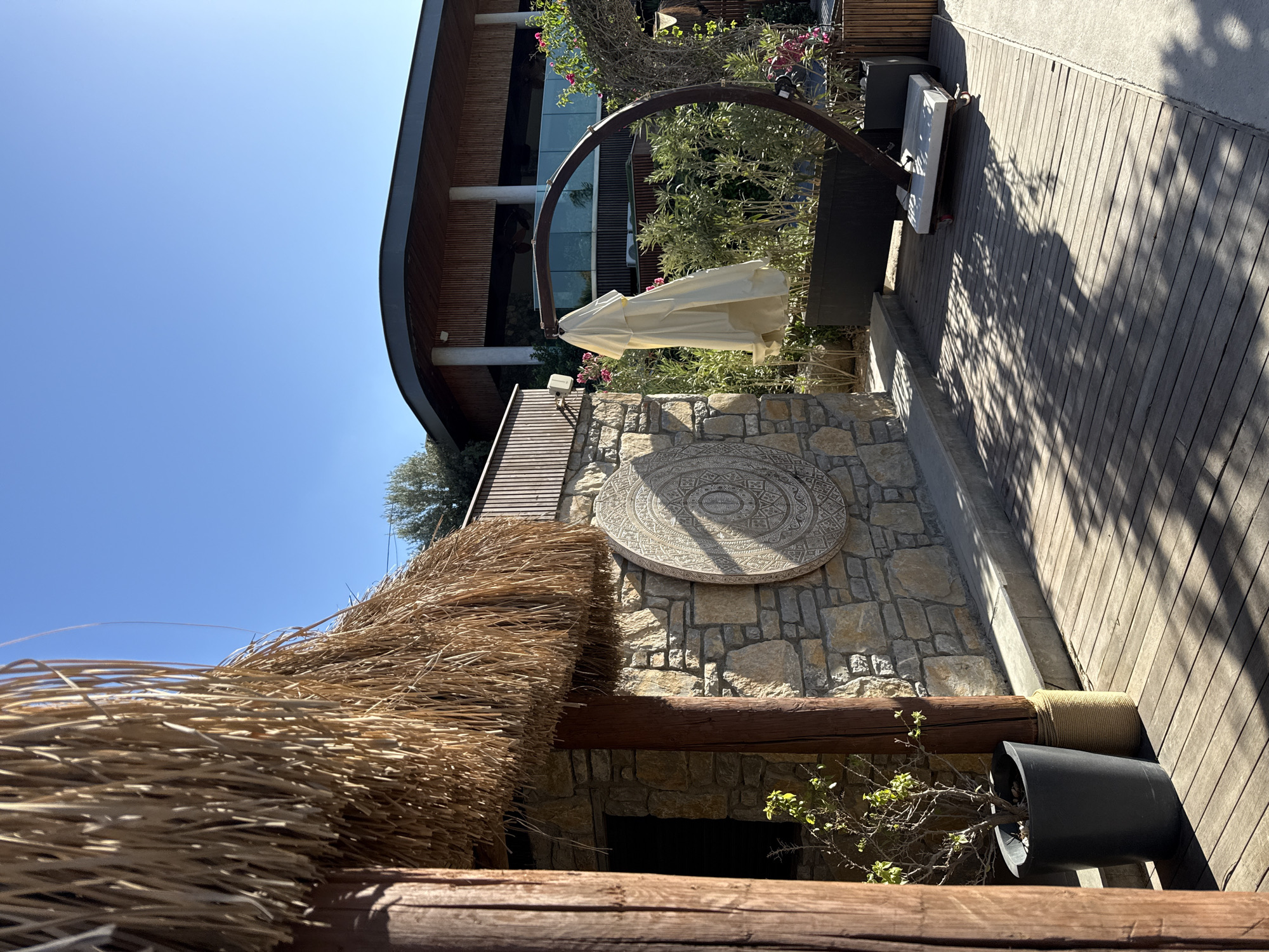 The image depicts an outdoor scene in Bodrum, Turkey, bathed in bright daylight under a clear blue sky. In the foreground, a thatched roof structure with wooden posts partially obscures the view. To the right, a wooden deck is laid out, casting dappled shadows from overhead foliage. Against a stone wall, a large circular decorative carving is visible. Further to the right, a modern building with a curved wooden facade and large windows stands in the background. Adjacent to the stone wall, a curved metal stand holds a cream-colored umbrella, folded and hanging. A white chaise lounge with cushions rests on the deck below the umbrella. Various potted plants and greenery surround the seating area, adding to the resort-like atmosphere. The overall setting suggests a tranquil, outdoor relaxation space.