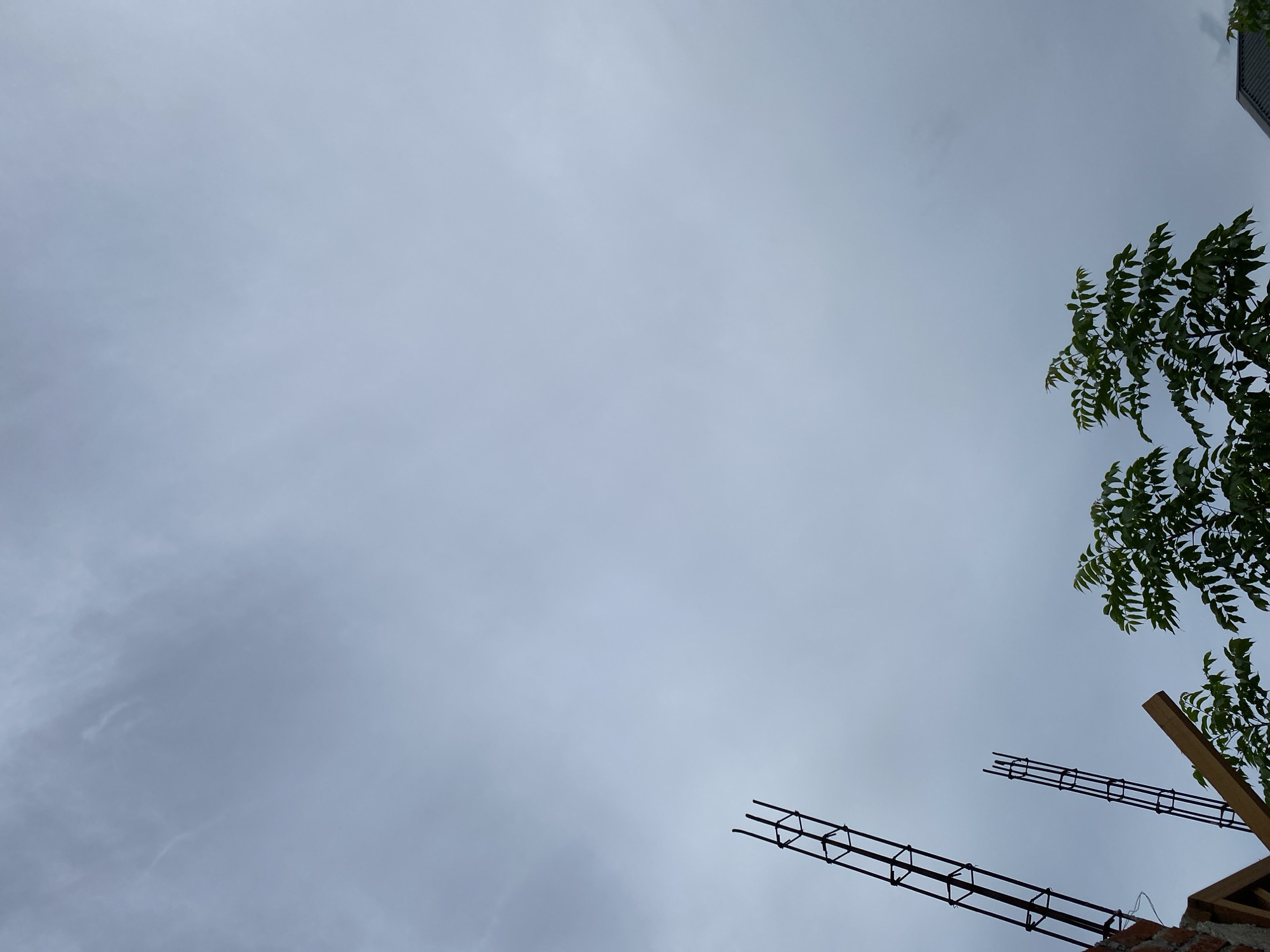 The image captures a cloudy sky with a construction site in the foreground. Two rebar structures, typical of building foundations, extend upwards. To the right, lush green trees with many leaves contrast with the muted sky. Part of a building's roof or structure is visible at the bottom right. The scene appears to be an outdoor environment in Banda Aceh, Indonesia, likely during daylight hours given the visibility.