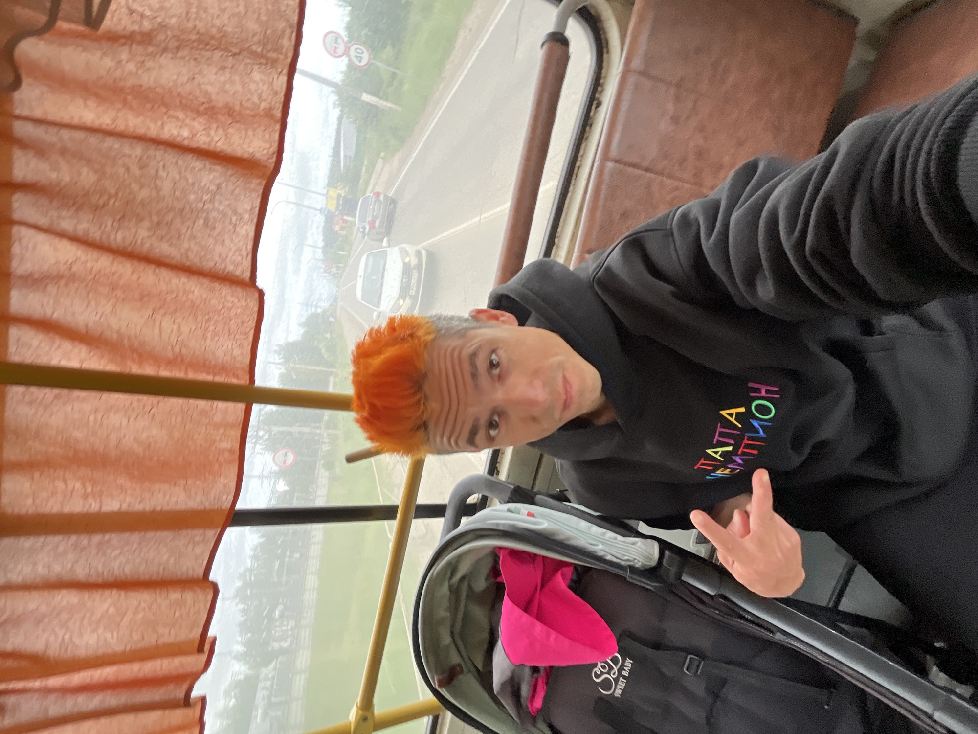 A light-skinned male with prominent orange, spiked hair is depicted taking a selfie inside a public transport vehicle in Chaikovsky, Russia. He wears a black hoodie with the text "ПАППА ЧЕМПИОН" (PAPA CHEMPION - "Dad Champion") embroidered in multi-colored letters across the chest, and black trousers. His left hand forms a "rock on" gesture. To his left, a grey and black baby stroller with a pink item inside is visible; the text "SWEET BABY" and an "S" logo are on the stroller's side pocket. The vehicle's interior features orange patterned curtains above the windows, a brown upholstered seat, and yellow and dark brown handrails. Through the window, a two-lane asphalt road is visible, with two white passenger cars traveling in the distance. Roadside elements include green foliage and three circular regulatory signs: a white sign with a red border and a horizontal black bar, a white sign with a red border and a vertical black bar, and a white sign with a red border displaying "40" (indicating a 40 km/h speed limit). The sky appears overcast.