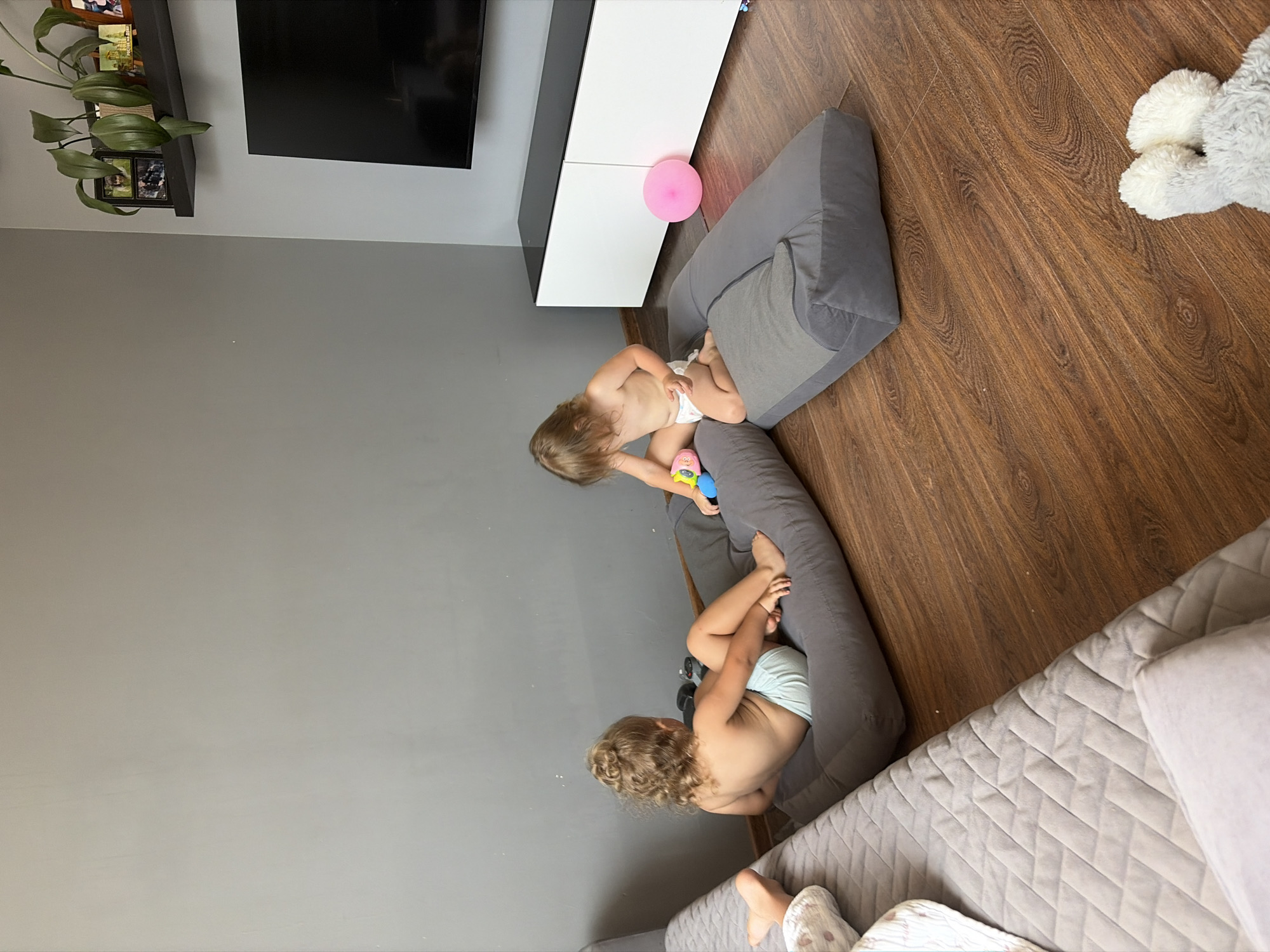 The image shows a living room scene. Two young children, a girl and a boy, are playing on large floor cushions. The girl on the left has curly blonde hair and is wearing a light blue diaper. She is sitting on a dark grey cushion and has toys in front of her. The boy on the right has straight blonde hair and is wearing a white diaper. He is also sitting on a dark grey cushion and is holding a colorful toy. The floor is made of dark wood laminate. To the left of the children is a grey quilted sofa. In the background, there is a grey wall, a white entertainment unit with a black top, and a large black television screen. A pink balloon is on the floor next to the entertainment unit. A shelf above the entertainment unit holds a potted plant and several framed pictures. The scene appears to be a home interior. The location is not specified in the image or the prompt to be Rostov-on-Don, Russia.