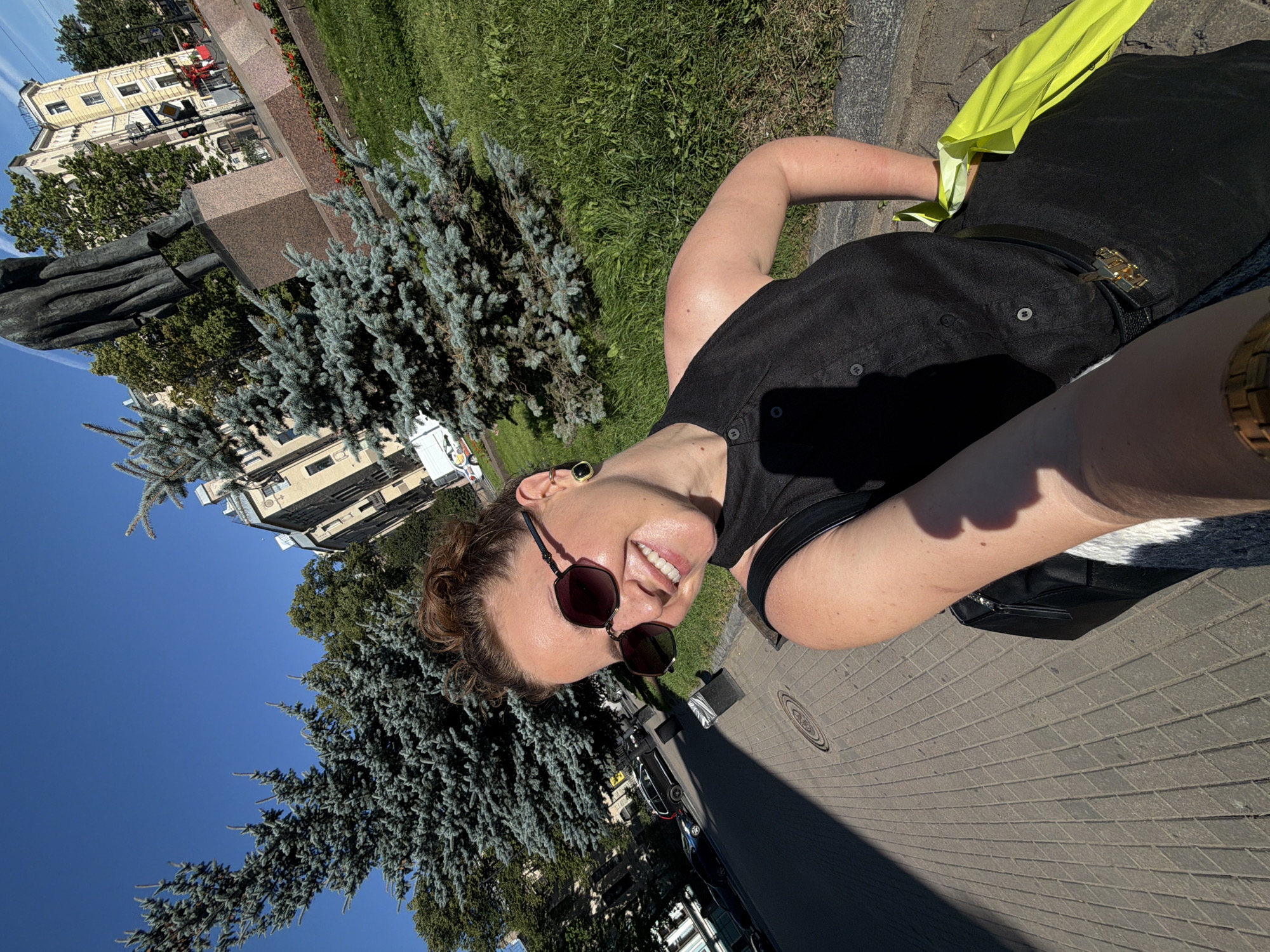 A woman is taking a selfie outdoors in Saint Petersburg, Russia, on a sunny day. She is wearing sunglasses, a black sleeveless dress, and a black belt with a gold buckle. A bright yellow bag is tucked into her belt. She has her hair pulled up in a bun and is smiling at the camera.
Behind her, there are several large coniferous trees, a statue of a person on a pedestal, and a light-colored building. The ground is paved with light-colored rectangular bricks. Cars are parked along the side of the road in the background. The sky is clear and blue. The overall scene suggests a pleasant day in a city park or square.