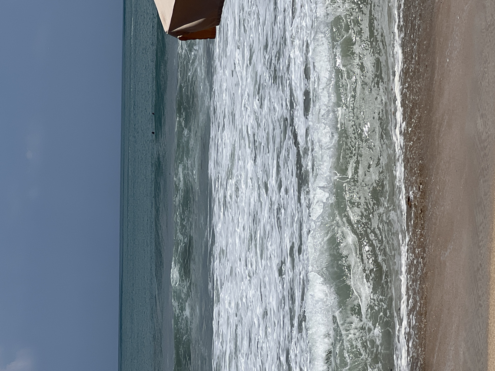 The image depicts a beach scene in Kuta, Indonesia, with a focus on the ocean and shoreline. In the foreground, the sandy beach meets the foamy edge of the water. Waves are rolling in, with the closest wave breaking and creating white foam. The ocean water is a greenish-blue hue, with several layers of waves visible stretching towards the horizon. Two figures, appearing to be swimming or surfing, are visible in the distance in the ocean. The sky is a hazy blue with some cloud cover. On the right side of the image, a portion of a light-colored beach umbrella is visible, suggesting a populated beach area. The overall impression is of a sunny day at the beach with moderate wave activity.