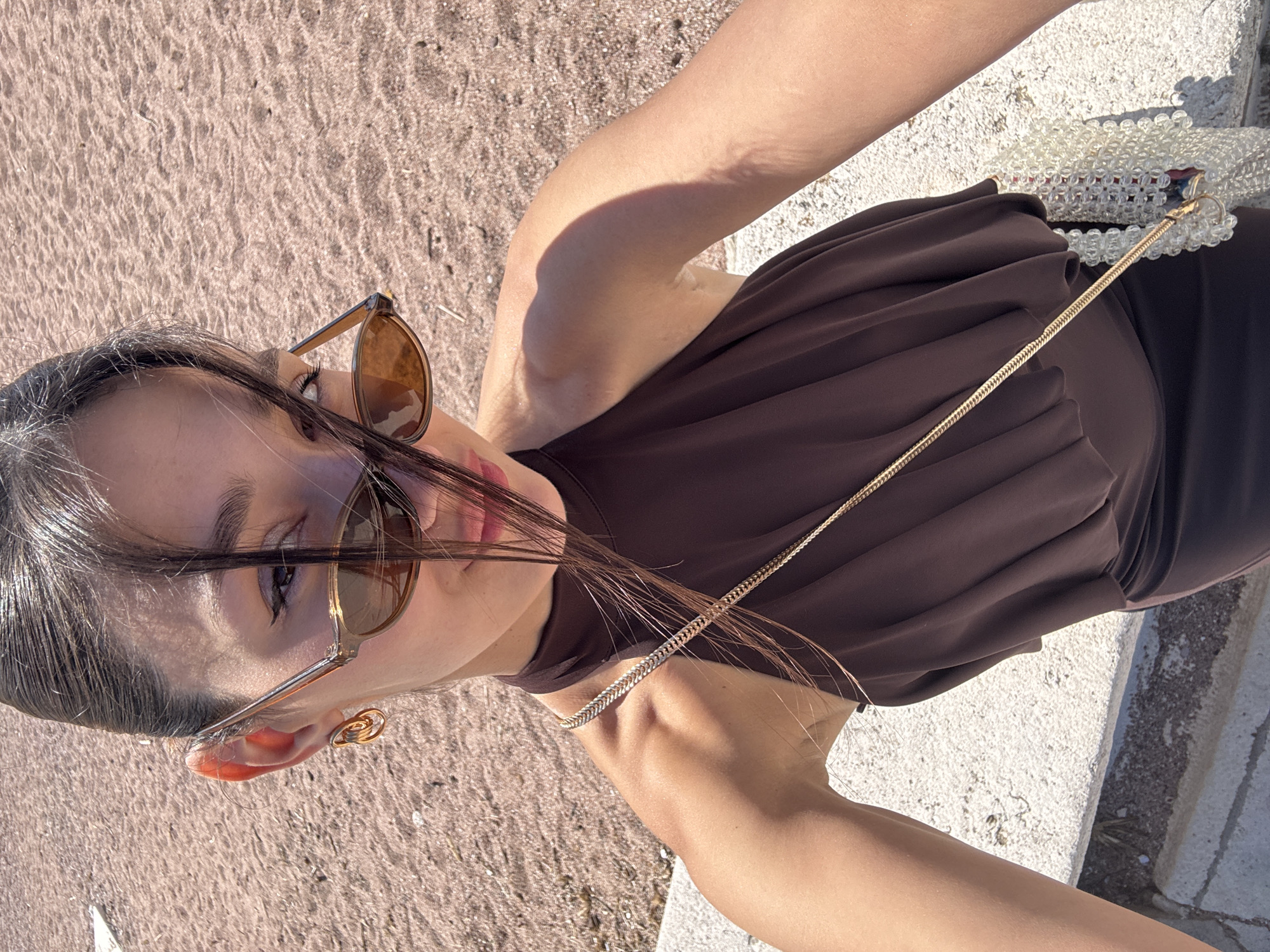 A woman with brown hair stands on a stone ledge near a sandy beach in Sveti Stefan, Montenegro. She is wearing sunglasses and a brown dress, and a purse with a beaded handle is visible on her right. The background features a beach and possibly other parts of Sveti Stefan.