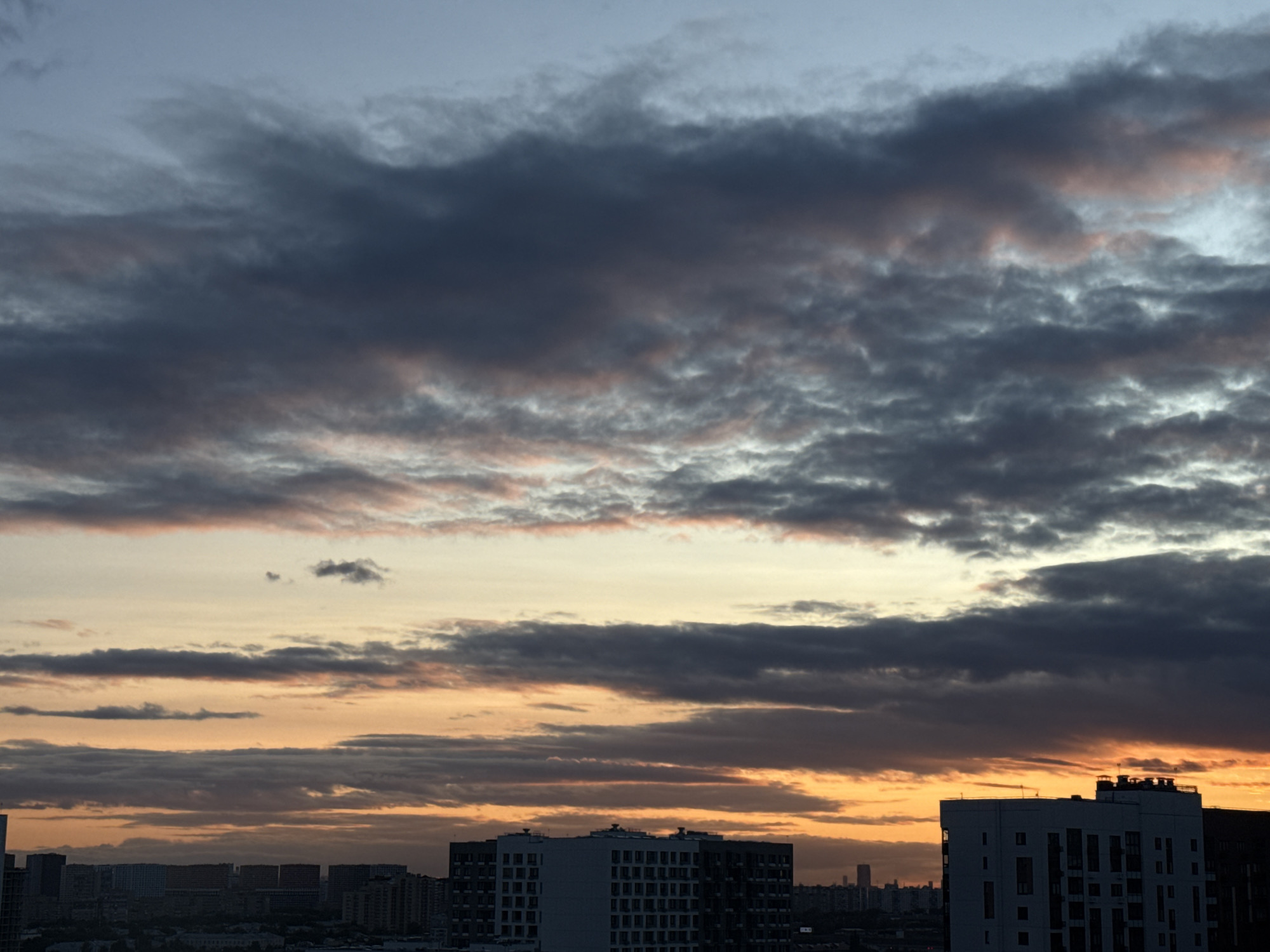 The image depicts a city skyline at dusk in Lyubertsy, Russia. The sky is filled with dramatic clouds, colored in shades of gray, dark blue, and orange from the setting sun. In the foreground, several modern apartment buildings rise above the city. The building on the right is particularly prominent, with a white facade and numerous windows. Other buildings, some taller and darker, are visible in the background, suggesting a dense urban environment. The overall impression is of a cloudy and colorful twilight over a residential area.