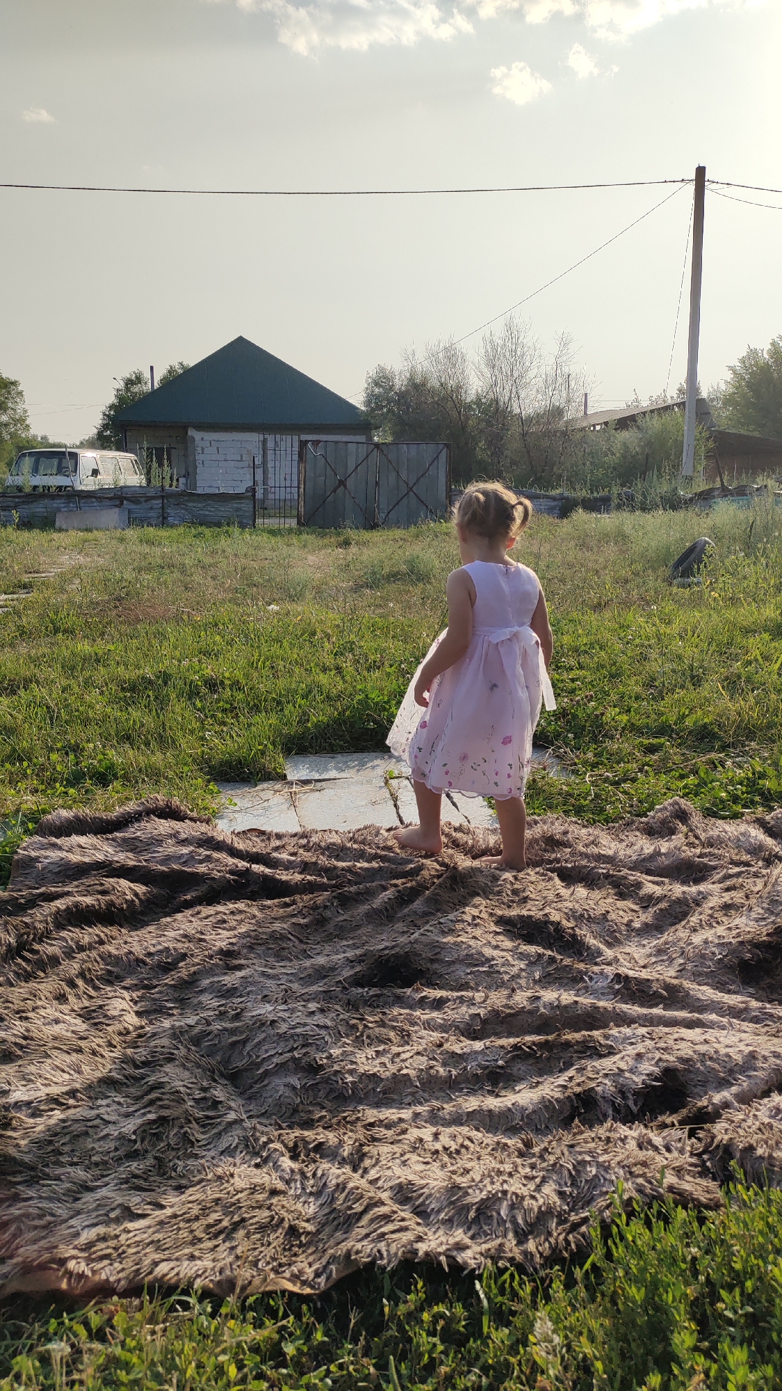 A young girl, wearing a light pink dress with floral patterns and her hair in two pigtails, stands barefoot on a shaggy rug. She is facing away from the camera, looking towards a somewhat dilapidated rural scene. In the background, there is a small, A-frame building with a green roof and white brick walls, possibly a shed or workshop, with a weathered metal gate in front of it. To the left of the building, a white van is parked. The area is overgrown with grass and weeds, suggesting a neglected or rural environment. A telephone pole with wires stretches across the sky, and a few wispy clouds are visible in the hazy sky. The lighting suggests it is late afternoon or early evening, with the sun casting long shadows. The overall mood is one of quiet, solitary play in a rustic setting.