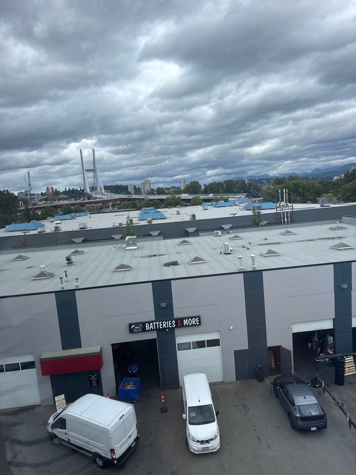 The image is an aerial view of a commercial building with "PHIL'S BATTERIES & MORE" signage. In front of the building, there are two vans and a car. The building is situated in Surrey, Canada, as evidenced by the distant view of the Alex Fraser Bridge. The sky is overcast, with dark clouds dominating the scene. The surrounding area appears to be an industrial or commercial zone with other buildings and infrastructure visible in the background. The overall atmosphere is grey and somewhat gloomy due to the weather conditions.