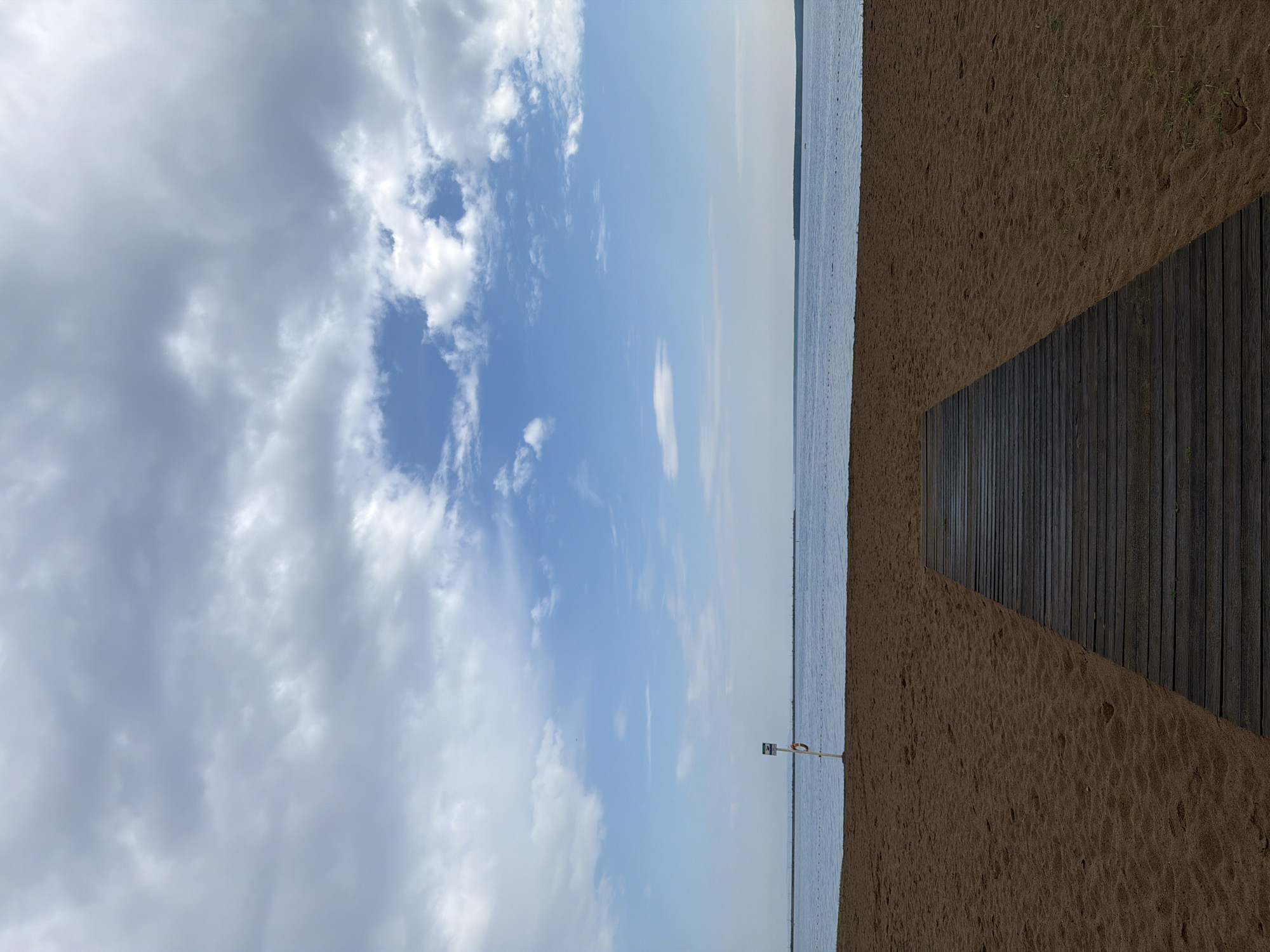 The image depicts a wooden boardwalk leading across a sandy beach towards a calm body of water under a cloudy sky. The scene is likely located on the Kama River, in the city of Chaikovsky, Russia, a region known for its beaches and recreational areas. In the distance, a distant tree line is visible across the water. Near the water's edge, a white pole with a sign and a lifebuoy is planted in the sand, suggesting a designated swimming or safety area. The sky is filled with a mix of light and dark clouds, with patches of blue sky visible. The sand is a warm brown color, with some faint footprints visible. The water is a tranquil blue-gray, with subtle ripples on its surface.