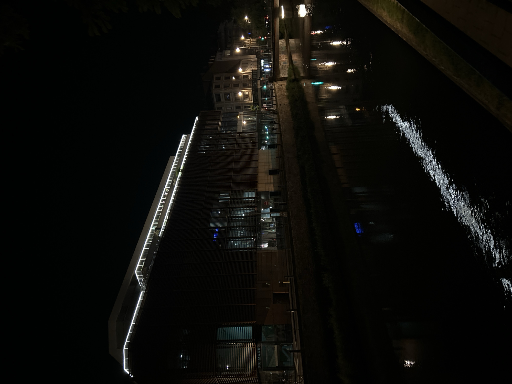 The image shows a night scene along the Ill River in Strasbourg, France. A modern building, illuminated with white lights along its edge, stands next to the river. Across the river, older buildings with lit windows can be seen. Reflections of lights shimmer on the water's surface. A portion of a bridge or walkway is visible in the upper right corner of the frame. A store called "Primark" is seen in the bottom part of the modern building.