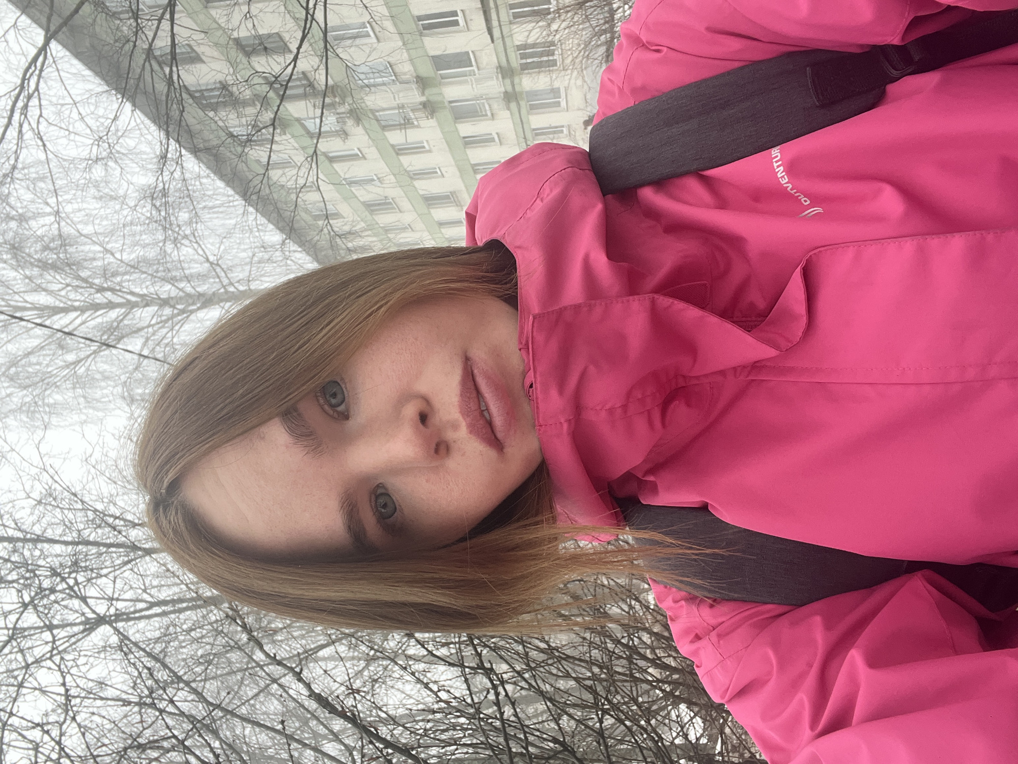 A young woman stands outdoors, wearing a bright pink hooded jacket and a dark grey backpack. The background reveals a high-rise building with multiple windows. Bare tree branches fill the lower part of the frame, suggesting a winter scene or a season with leafless trees. The location is identifiable as Chaikovsky, Russia, based on the architectural style of the building and general environment.