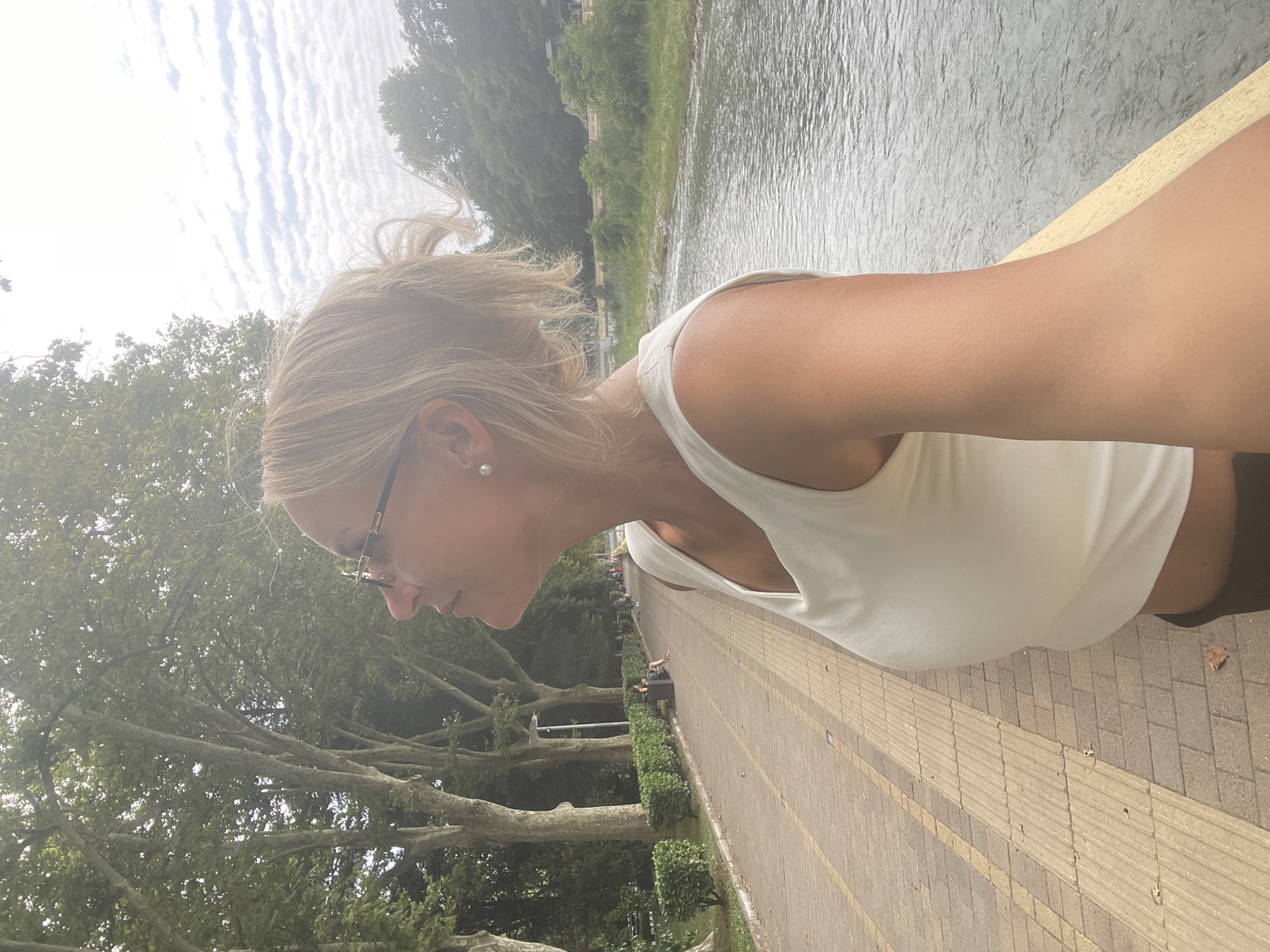 A woman with blonde hair pulled back and wearing glasses and a white top is standing on a paved walkway next to a river. The walkway has a pattern of yellow and brown tiles. Trees line the left side of the walkway, and in the distance, a few people are visible sitting on a bench and walking. The river on the right is calm with ripples. The sky is cloudy. This scene is likely in the city of Sochi, Russia, given the context provided.