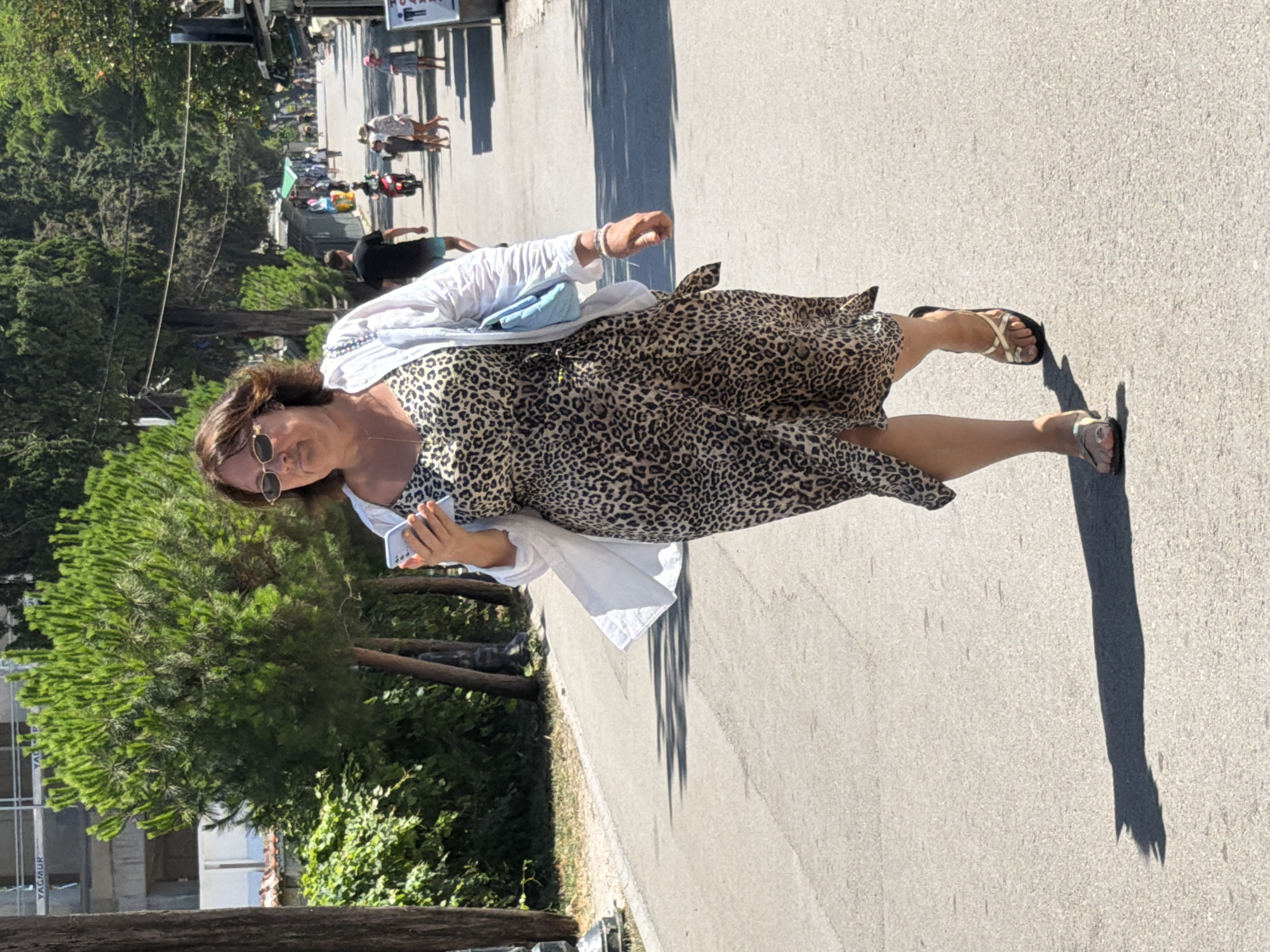 A woman with brown hair and sunglasses is walking down an asphalt road in Budva, Montenegro. She is wearing a leopard-print dress, a white open shirt, and gold sandals. She is holding a light blue handbag and a smartphone in her hands. In the background, there are trees, buildings under construction, and other people walking and riding a scooter. The scene is illuminated by bright sunlight, casting shadows on the road.