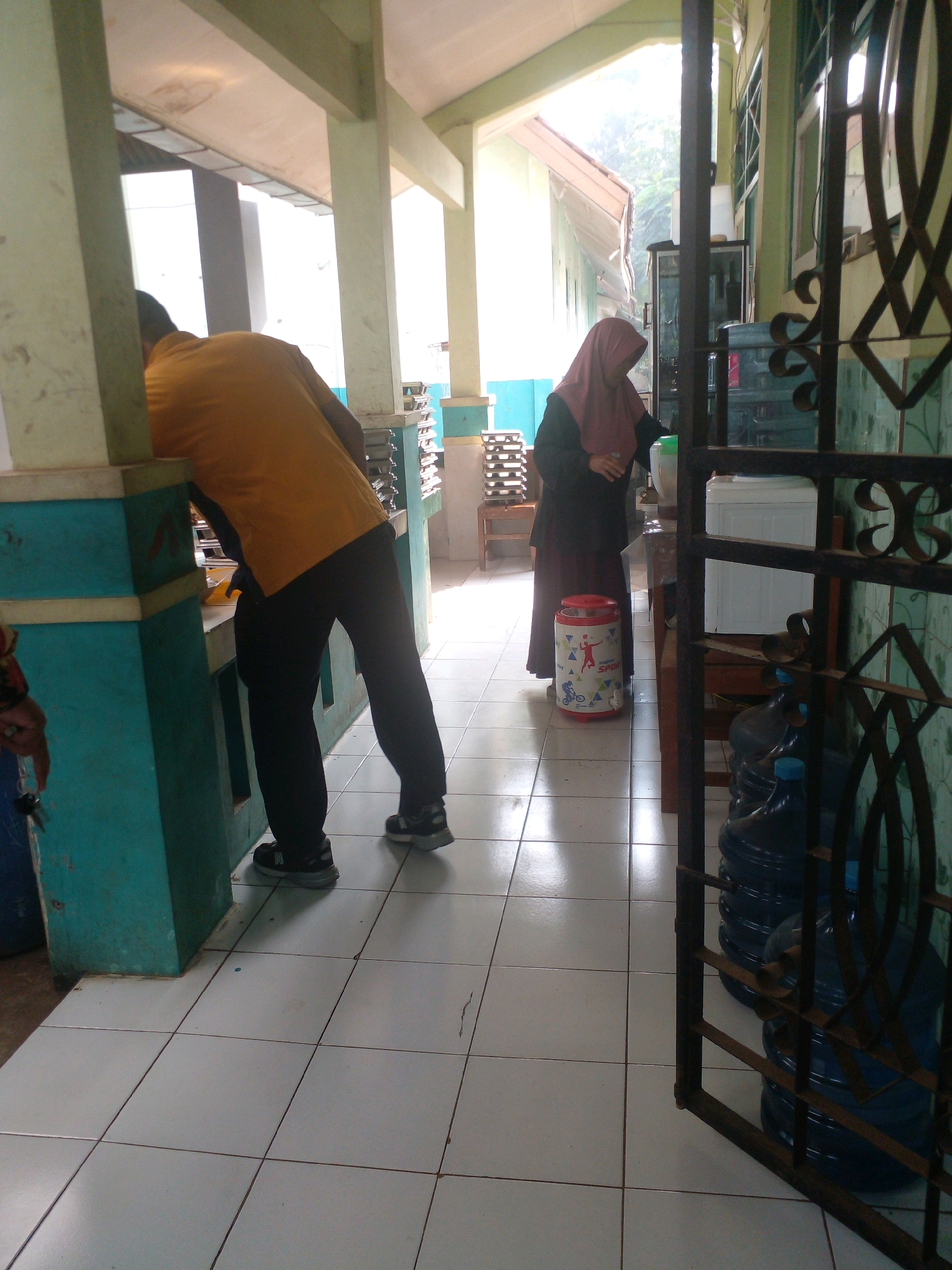 The image depicts a hallway or covered outdoor area with tiled flooring. On the left, a person wearing an orange shirt and black pants is partially visible, leaning over a counter. To the right, a person wearing a dark hijab and a dark robe is standing near a water dispenser. Several large water bottles are stacked to the right of the water dispenser, partially obscured by a black metal gate. The environment appears to be a public space, possibly a school or community center, with light filtering in from an open area. The specific location within Banyuputih, Indonesia, cannot be determined from the visual information provided.