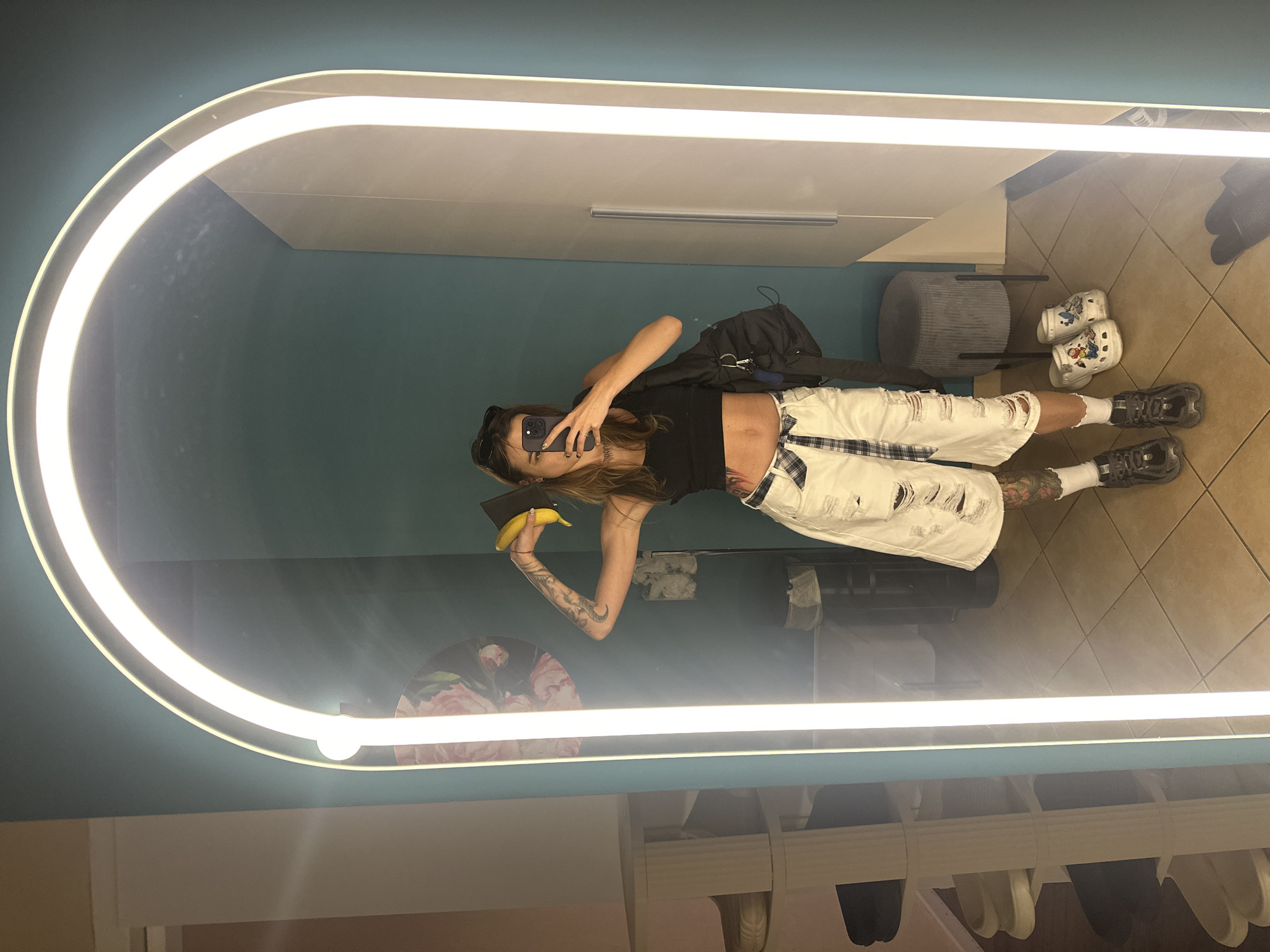 A full-length mirror with an arched LED light frame dominates the foreground of the image. A woman is standing in front of the mirror, taking a selfie with her phone. She is wearing a black crop top, white ripped denim shorts, and dark sneakers. A black backpack is slung over her left shoulder. She is holding a banana in her right hand. The background appears to be an indoor changing room or dressing area with teal walls. To the left of the mirror, a white multi-tiered shelf is visible, holding several pairs of shoes. To the right, a grey ottoman and a white cabinet are present. The floor is tiled. The woman has tattoos on her arms and legs. The scene does not provide any specific indicators of its location in Moscow, Russia.