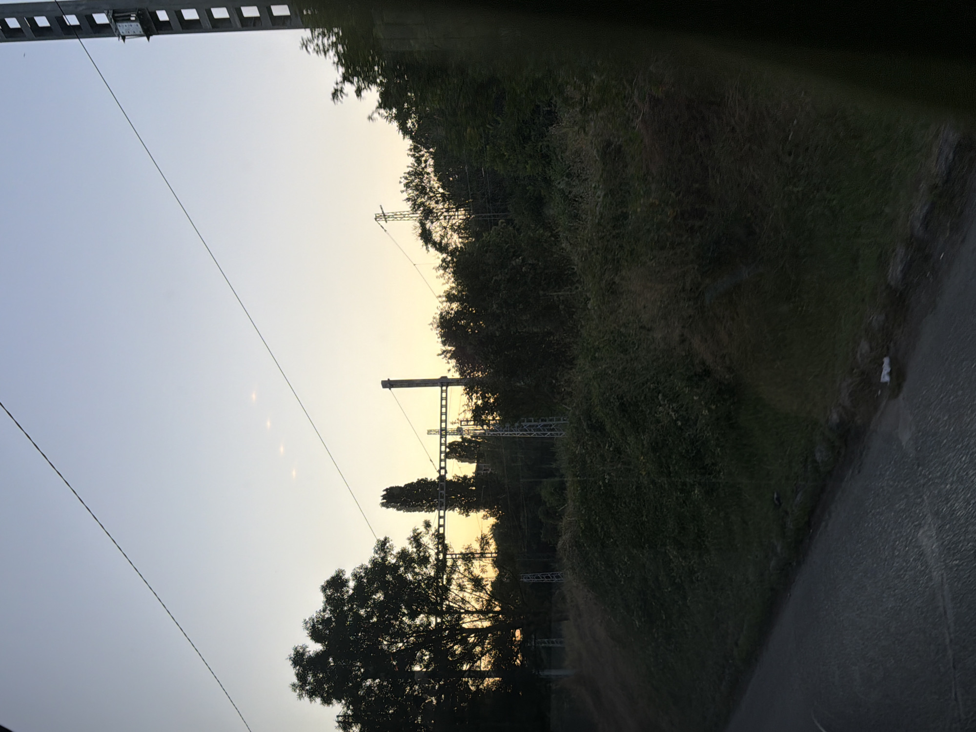The image captures a view of overhead power lines and trees at sunset, with a clear sky transitioning to a pale yellow near the horizon. A tall, lattice-like electrical pylon stands on the right side of the frame, its structure outlined against the sky. In the foreground, a paved surface, likely a road or path, runs diagonally from the bottom left corner. The landscape beyond the road is densely covered with green vegetation and trees, some of which are silhouetted against the twilight sky. The overall scene suggests a rural or semi-urban environment on the outskirts of Sofia, Bulgaria, given the presence of power infrastructure and natural elements typical of such areas. The lighting indicates it is either early morning or late evening. No people or specific actions are depicted.