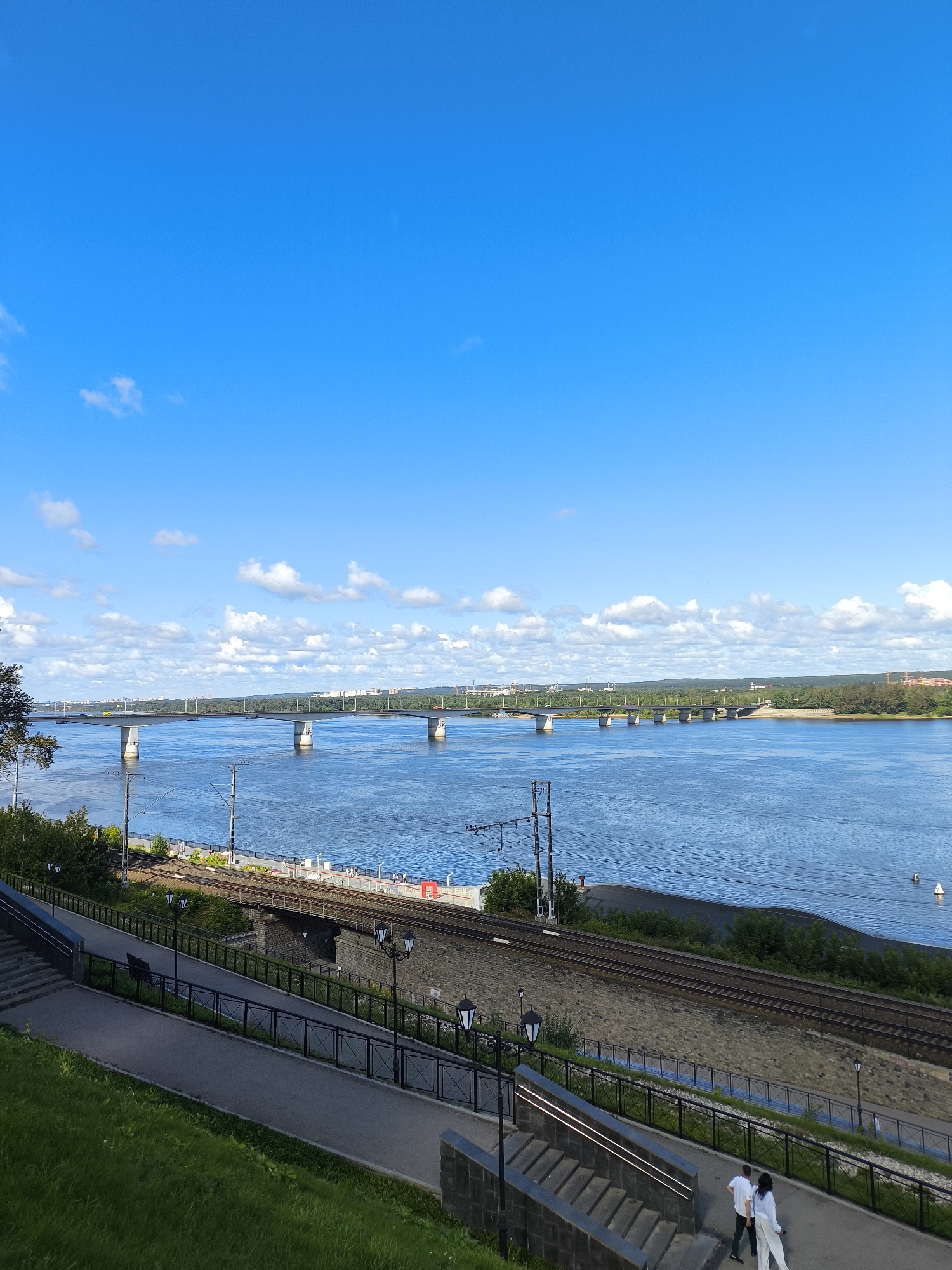 The image shows a view of the Kama River and a bridge in Perm, Russia, on a sunny day with blue skies and scattered white clouds. In the foreground, a grassy slope leads to a paved walkway with stairs, lampposts, and a black metal fence. A set of train tracks runs parallel to the walkway and river. Two people, a man and a woman, both dressed in white, are walking away from the camera on the walkway. The bridge, which appears to be a railway bridge, spans the river in the middle ground. The far bank of the river is lined with trees and buildings.