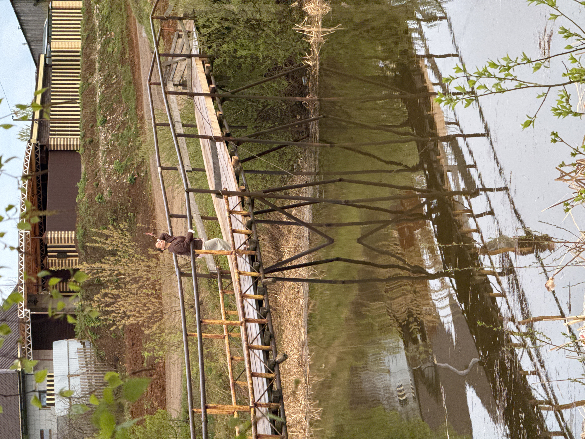 The image shows a woman sitting on a wooden and metal pier that extends into a body of water, likely a river or a lake. The pier is old and appears to be rusty in places. The water reflects the structure of the pier and surrounding vegetation, including trees and tall grasses along the shoreline. The woman is wearing a brown jacket and light-colored pants, and she is positioned towards the end of the pier, appearing relaxed.
In the background, a building with striped exterior walls is visible among the foliage. Adjacent to this building, another structure with a light-colored exterior and gabled roof is partially visible. The scene is set during daylight, and the lighting suggests it is either early morning or late afternoon. The location is identified as Chastyye, Russia, based on the prompt.