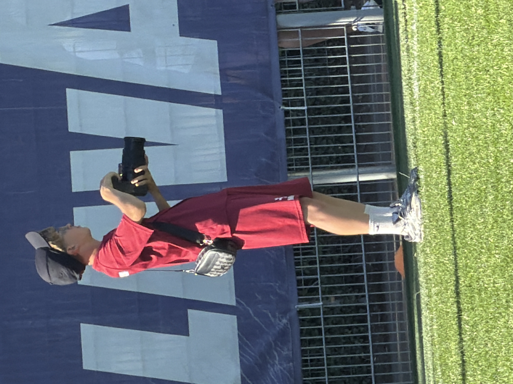 A person wearing a red shirt and shorts is standing by a fence, taking a photo with a camera in Fentange, Luxembourg. They are wearing a dark-colored hat, socks, and athletic shoes. A small bag is hanging from their shoulder. The backdrop includes a large blue sign with white geometric shapes and a green grassy area. The scene is outdoors and appears to be during daylight hours.