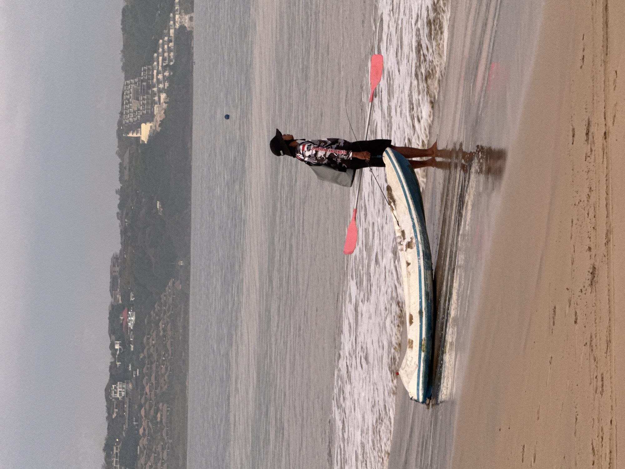 The media file captures a coastal scene in Kuta, Indonesia, featuring a person and a kayak on a beach. The image is oriented vertically, but depicts a horizontally expansive view of the ocean and shoreline.
In the foreground, a single individual stands in the shallow, foamy water where gentle waves meet the sandy shore. The person is wearing a black hat, a long-sleeved shirt with a black, white, and grey camouflage-like pattern, and dark shorts. They are partially turned away from the camera, looking towards the vast ocean. Text on their shirt reads "BUMBARAN ANGLER".
Next to the person, a white kayak or small outrigger canoe with blue stripes rests partially in the water and on the wet sand. Two paddles with distinct red blades are visible, one propped against the kayak and another near the person. The beach itself is a light tan color, with wet areas reflecting the sky.
The ocean stretches into the distance, appearing calm with small, breaking waves near the shore. A small, dark blue buoy or floating object is visible further out in the water. The background reveals a lush, green hilly coastline densely populated with numerous light-colored buildings, indicative of resorts or residential areas. The sky is hazy or overcast, contributing to soft, diffused lighting. The scene suggests a quiet moment during the daytime, possibly before or after a kayaking activity, with the individual observing the seascape.