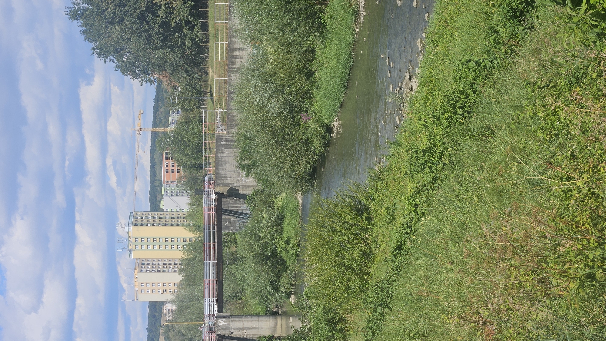 The image depicts a river flowing through a semi-urban area, likely in Prešov, Slovakia, given the context of other images from this set. A concrete bridge spans the river, supporting a road with railing. On the left bank of the river, lush green vegetation and trees are prominent. In the background, several apartment buildings, characterized by their colorful facades and multiple stories, are visible, suggesting a residential area. A construction crane stands tall amongst the buildings, indicating ongoing development. The sky is partly cloudy, with a mix of blue sky and white clouds. The overall scene captures a blend of natural elements, urban infrastructure, and signs of construction.