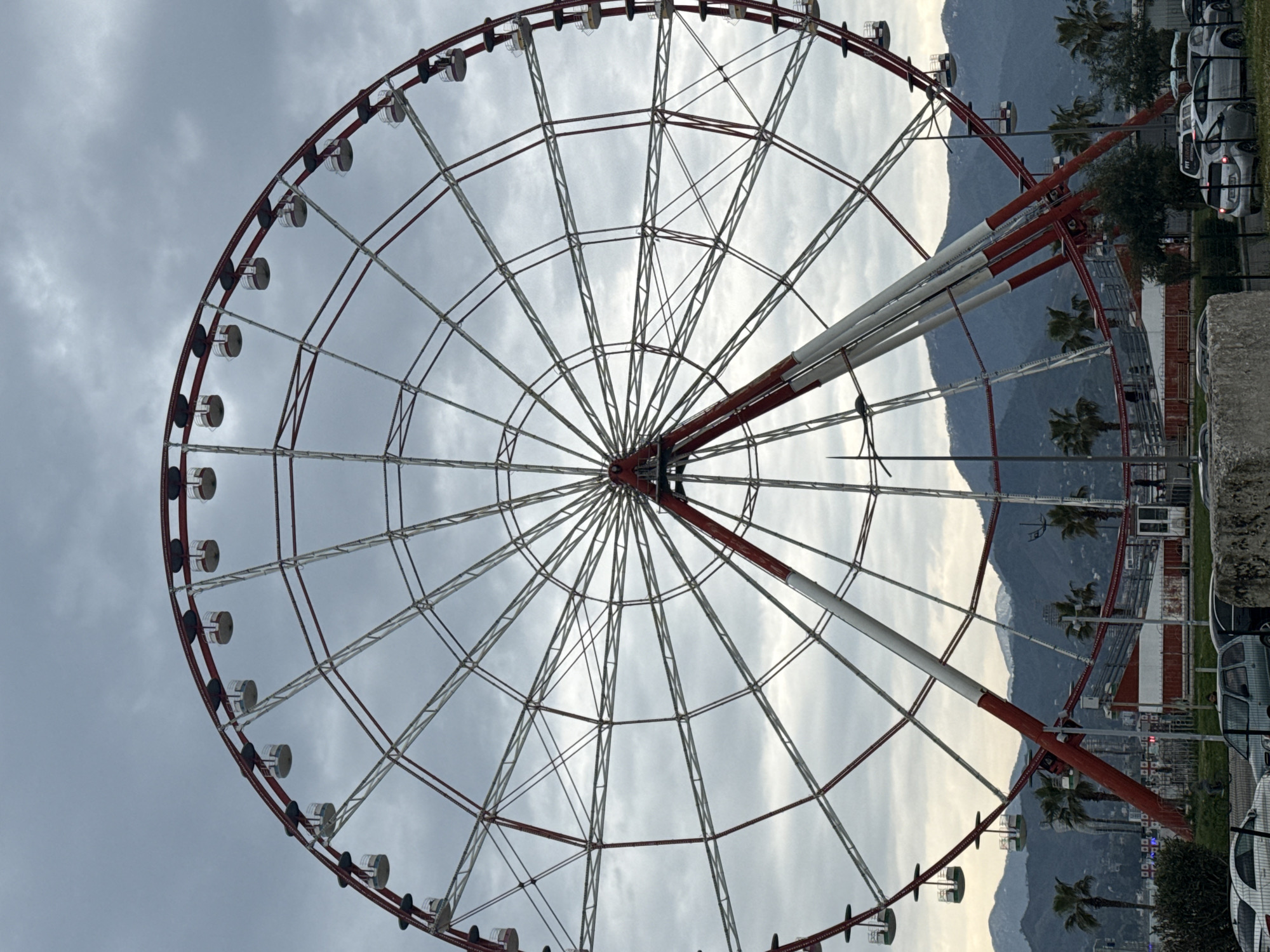 The image shows a Ferris wheel located in Batumi, Georgia. The wheel has a red and white frame and is surrounded by a cloudy sky. Mountains and palm trees are visible in the background, and the Ferris wheel is situated near buildings, trees and parked cars.