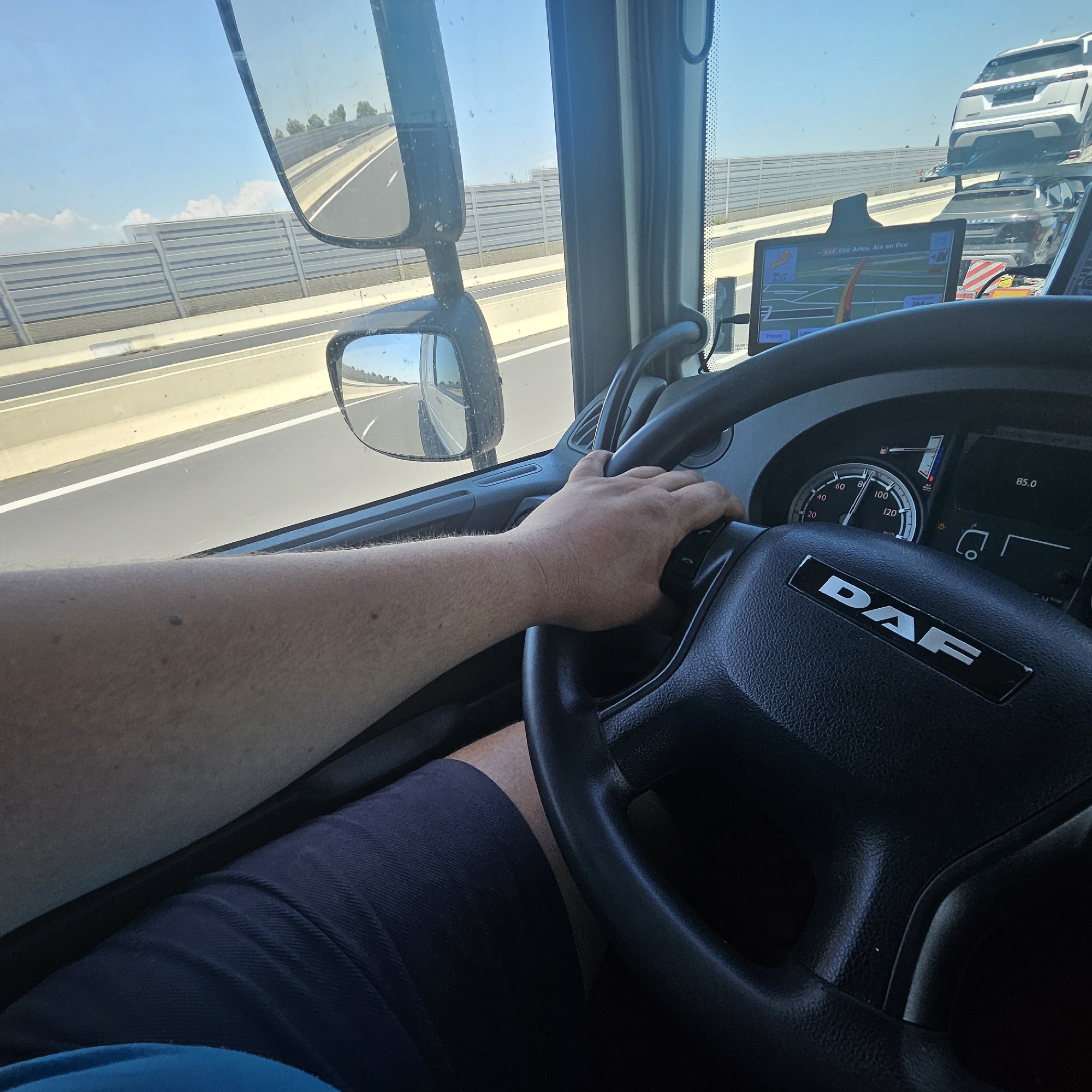 The image, taken from within the cabin of a DAF truck, reveals a driver's-eye-view of a highway, identified by the navigation system as "A54 E80 Arles, Aix en Pce". The driver's left hand is visible on the steering wheel. The truck's dashboard shows a speed of 85 km/h. In the distance, a vehicle transport truck is seen carrying JASCOO-branded cars. The reflection in the side mirror and the view out of the windshield show a multi-lane highway with concrete barriers. The image suggests the truck is in transit, presumably somewhere along the A54 or E80 motorway in France. It is not possible to confirm that this is specifically in the city of Lattes, France.