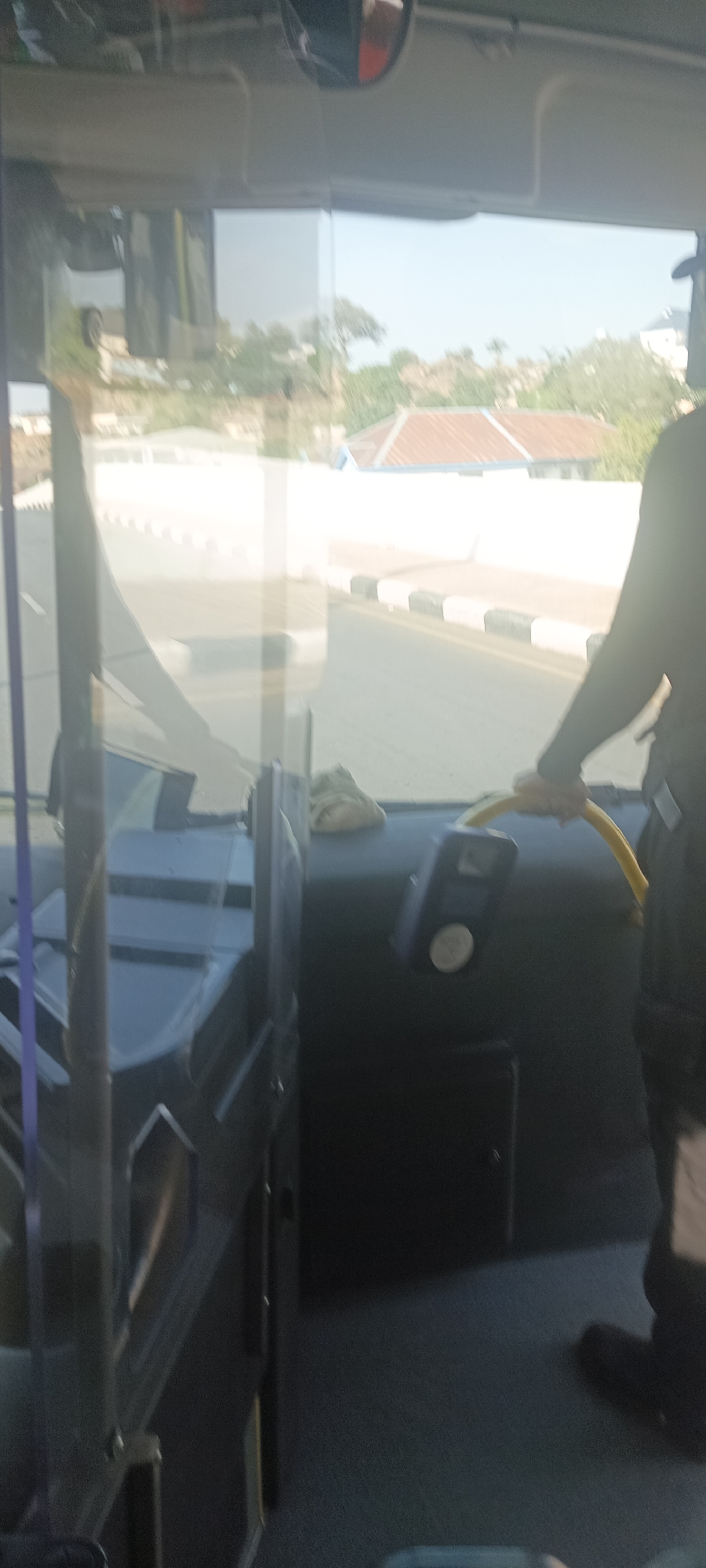 The image depicts the interior of a bus, presumably in motion, near what appears to be a residential area. A driver, partially visible on the right, is holding the yellow steering wheel. The road outside features a white wall, a house with a red roof. The location is Jos, Nigeria based on the visual cues in the photograph.