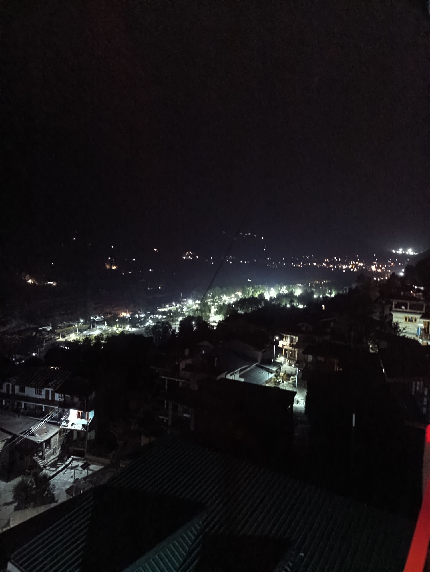 The image shows a nighttime view of Vashisht, India, featuring a landscape dotted with numerous lights from homes and streetlights. The scene captures a broad perspective of the town, extending into the background where more distant lights are visible against a dark sky. The foreground is dominated by the roofs of several buildings, some with visible windows and architectural details, while the midground displays a cluster of structures illuminated by artificial light, hinting at residential and possibly commercial activity. The overall impression is one of a town settling in for the night, with its lights creating a warm glow against the darkness.