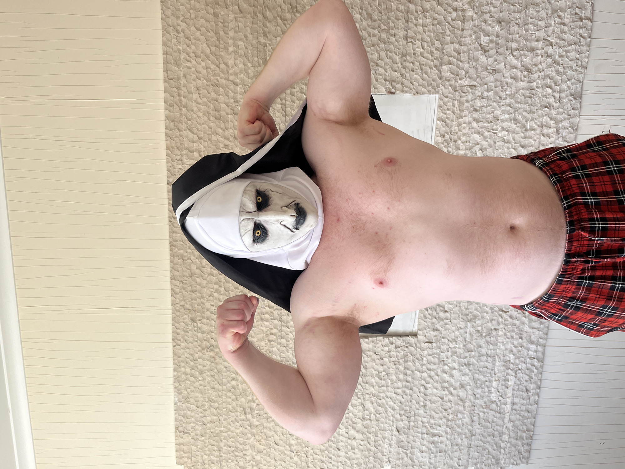 A shirtless man wearing a "The Nun" mask and red plaid boxer shorts stands indoors. He is flexing his biceps. The background consists of cream-colored walls and a light-colored textured rug. There is no indication of the location being in the city of Ореховка, Russia.