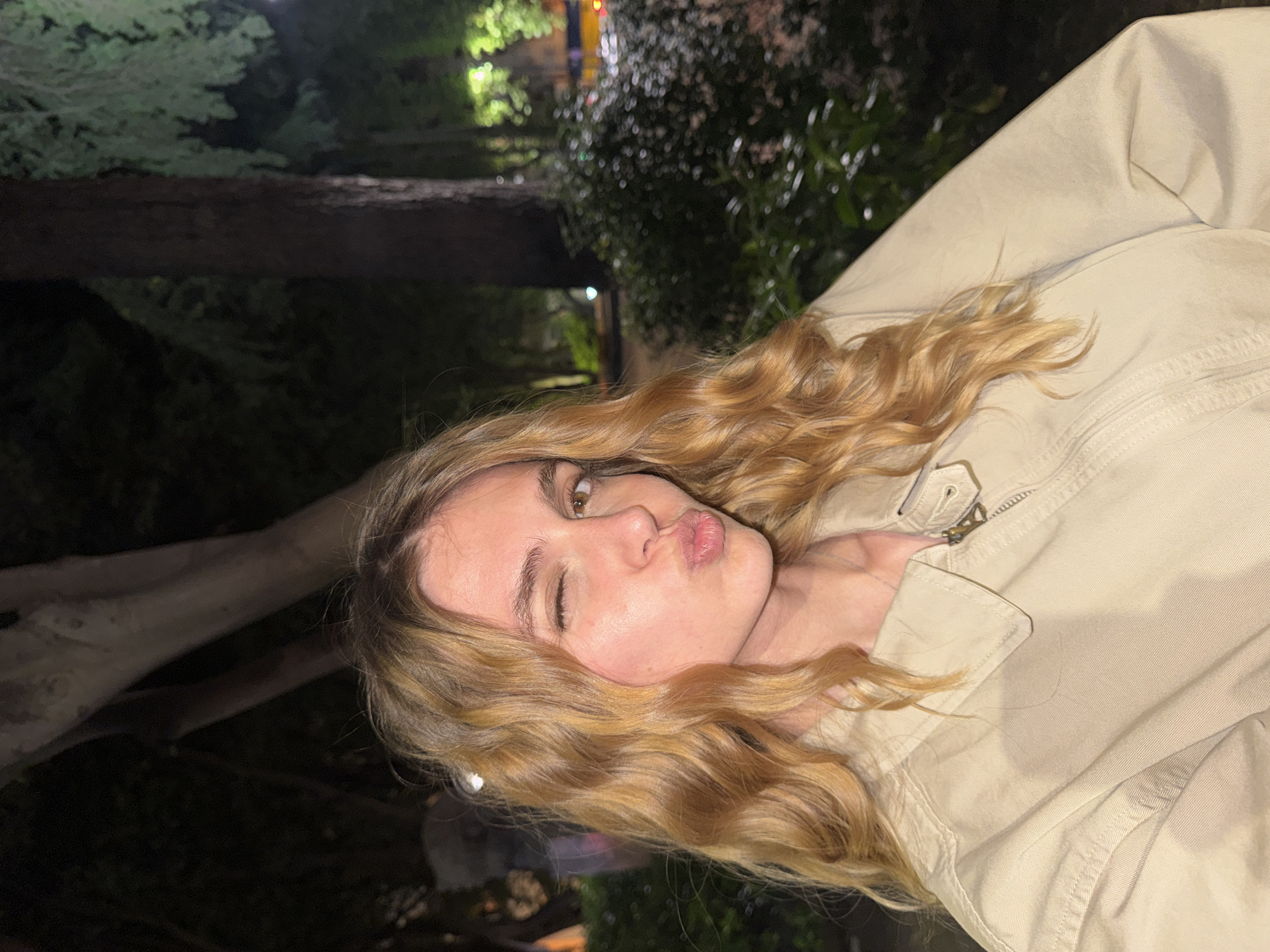 A woman with wavy blonde hair wears a beige jacket and winks with pursed lips. The background is blurred with trees and lights, potentially indicating an outdoor setting at night. The location is Tbilisi, Georgia.