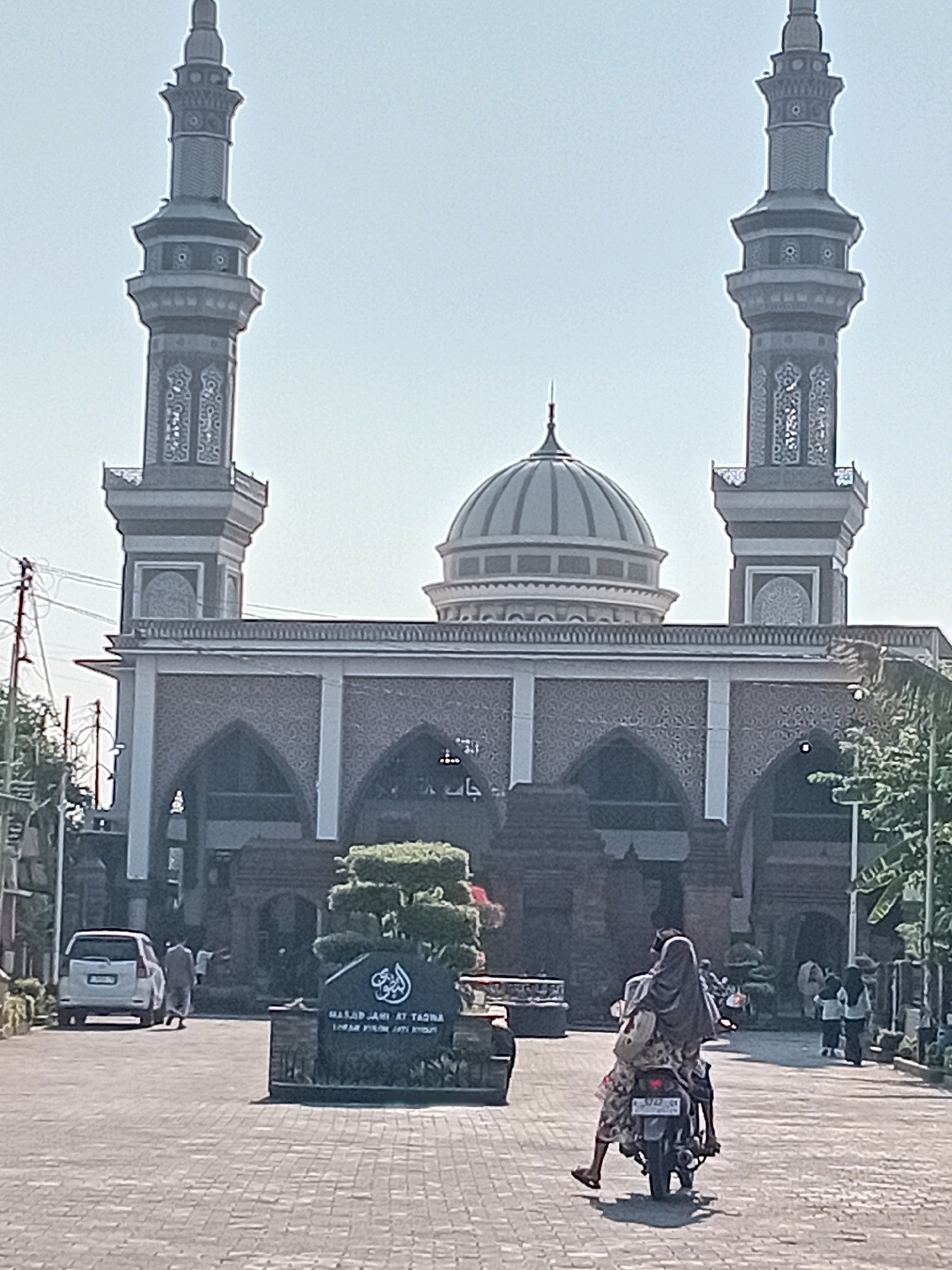 The image captures the Masjid Jami' At Taqwa in Loram Kulon, Indonesia. The mosque features a prominent dome and two tall minarets. In the foreground, a person is riding on the back of a motorcycle, facing away from the camera, wearing a headscarf and a patterned outfit. Another person is driving the motorcycle. To the left, a white van is parked, and several people are walking in the background near the mosque entrance. A stone sign with Arabic calligraphy and text identifying the mosque is situated in the middle ground. The ground is paved with cobblestones, and the scene is illuminated by natural daylight under a clear sky.