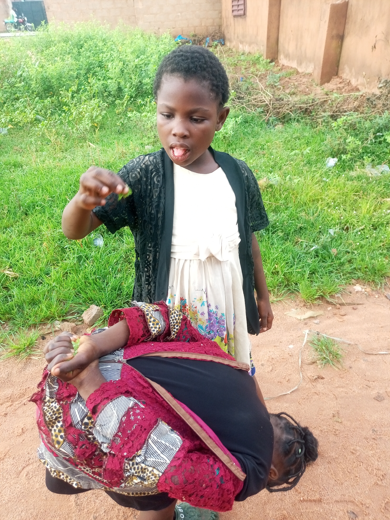 In an outdoor scene, likely in Tempure, Nigeria, two young Black girls are the central subjects. One girl, standing, wears a white dress with a floral print and a black lace overlay, and is holding something small and green in her right hand, bringing it to her mouth. She appears to be looking downwards. The second girl is lying on her side on sandy ground, her head bent towards the dirt. She is wearing a red garment with a patterned overlay and a black top. The environment is characterized by dry, sandy soil, patches of green grass, and a brick building with a tiled roof in the background. A motorcycle is partially visible to the far left. The lighting suggests daytime.