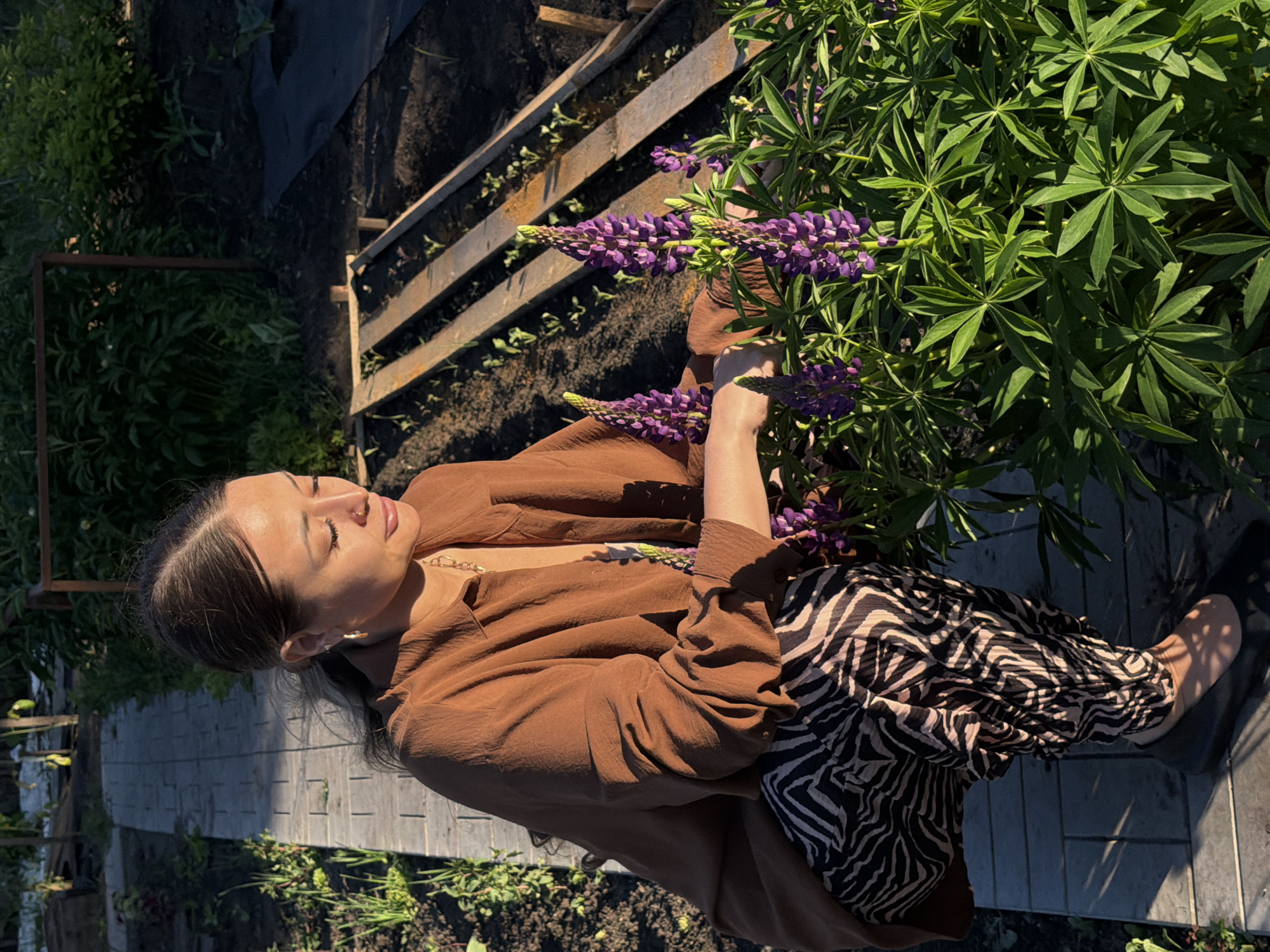 A woman is crouching down next to a cluster of purple lupine flowers in a garden. She is wearing a brown oversized shirt and patterned pants. The garden has a stone pathway and raised garden beds with young plants. The scene is lit by the warm light of the sun, casting shadows across the pathway. The location is identified as Kansk, Russia.