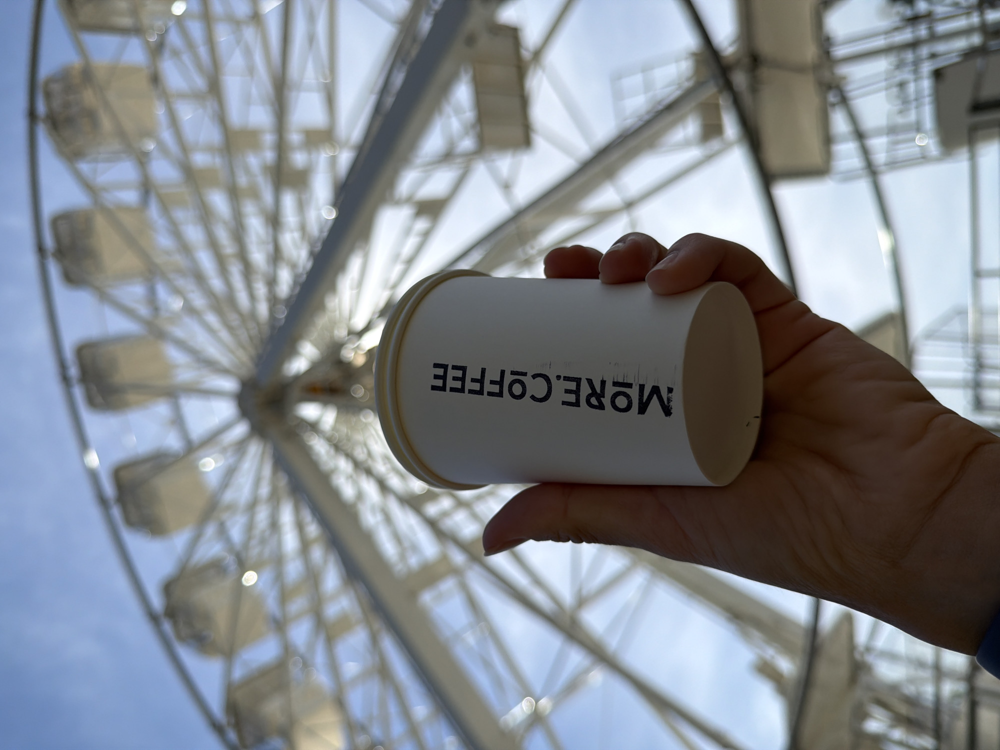 A low-angle shot shows a hand holding a white paper coffee cup with "MORE.COFFEE" printed in black, oriented toward the viewer. The coffee cup is slightly blurred, and the hand holding it is somewhat out of focus. In the background, a large Ferris wheel extends toward the sky, which is partly visible in the gaps between the wheel's structure. The Ferris wheel is out of focus, and the scene appears to be taken during daylight hours. The Ferris wheel is located in the city of Budva, Montenegro.