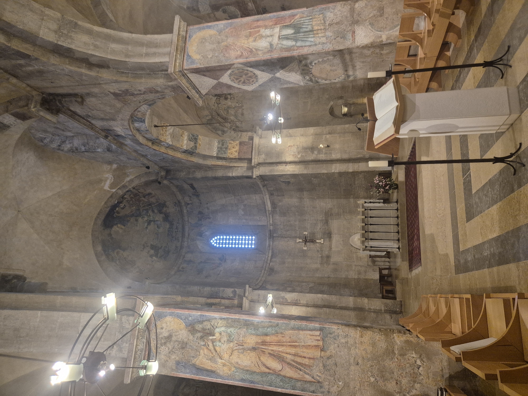 The image shows the interior of a stone church. In the foreground, there are rows of wooden benches. To the right, a white lectern stands with an open book on it. Two black candelabra with white candles flank the lectern. In the mid-ground, there is an altar with a white cloth, and a red rug on the floor. A large stained-glass window with a blue diamond pattern is visible on the back wall, with a cross hanging below it. There are frescoes and murals on the walls and pillars of the church, depicting religious figures and patterns. The lighting suggests it is daytime.