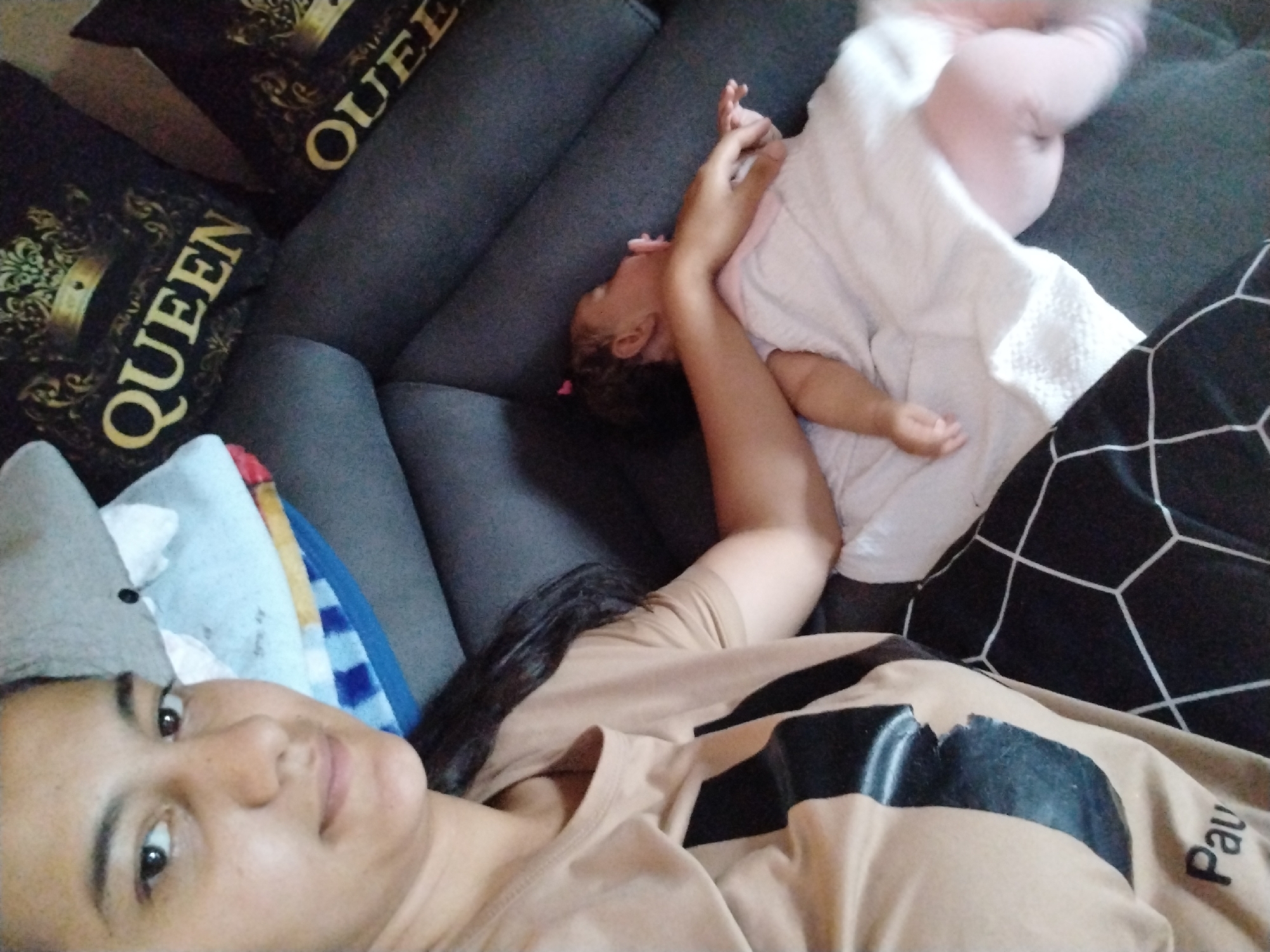An overhead, slightly angled shot depicts a woman and a child resting on a dark gray upholstered sofa in an indoor setting within the city of Unknown, Unknown. The woman, positioned in the foreground on the left, has dark hair and a light brown complexion, and is wearing a tan t-shirt with black graphic elements and the partially visible word "Paul" printed on it. She is looking directly towards the camera. To her right and slightly behind her, a young child, possibly an infant or toddler, with dark curly hair and a small pink hair clip, lies on the sofa, partially covered by a white textured blanket or garment, with pink clothing visible underneath. The child's arm is raised, and the face is visible in profile, turned away from the camera. Two black decorative pillows, each featuring a gold crown and the word "QUEEN," are visible on the sofa to the left of the child. A blue and white striped fabric is partially visible behind the woman's head, and a black fabric with a white geometric grid pattern is visible on the far right edge of the frame.