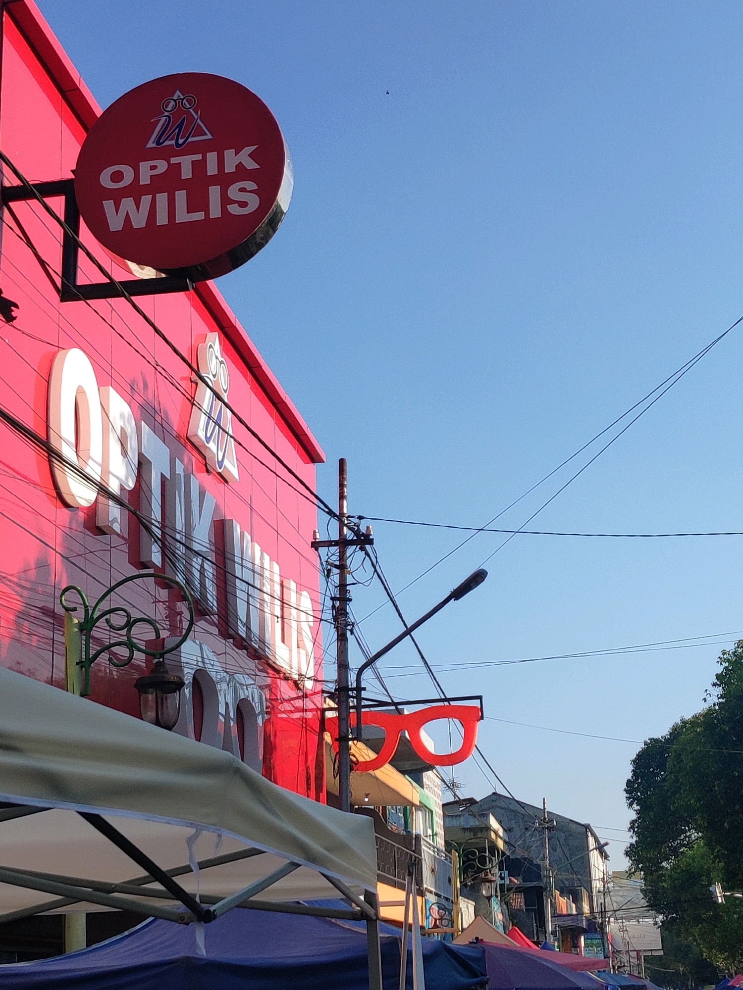 A street-level outdoor view captures a bright red building with large white lettering spelling out "OPTIK WILIS" on its facade. A circular red sign with "W" and glasses logo above "OPTIK WILIS" is mounted on the upper left of the building. A large red cutout of glasses hangs from a pole in front of the building. A street light is visible on the right. The sky is clear and blue. In the foreground, part of a canopy tent is visible. The scene is located in Kebonagung, Indonesia.