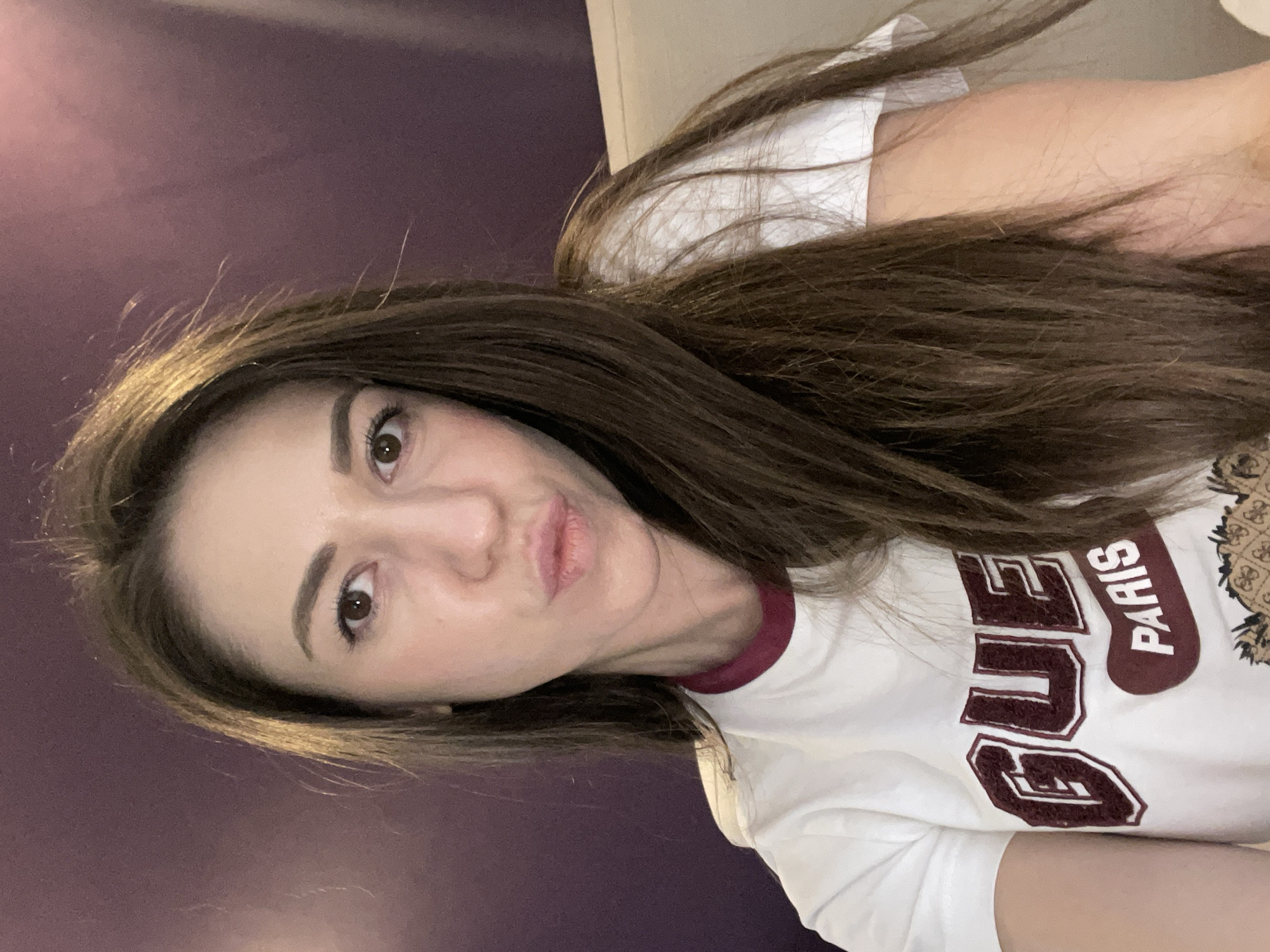 A close-up selfie shows a woman with long, straight, dark brown hair, which falls over her shoulders. She is looking directly at the camera with dark brown eyes and a subtle pout or "duck face" expression, tilting her head slightly. Her eyebrows are well-defined.
She is wearing a white short-sleeved t-shirt with a maroon-colored trim around the neckline. The t-shirt features prominent design elements, including large, maroon-colored embroidered letters that spell "GUE". Below and to the right of these letters, the word "PARIS" is visible, oriented vertically on a maroon patch. Adjacent to this text, there is a patterned emblem in shades of brown and black, possibly a brand logo.
The background consists of a plain, dark purple wall, indicating an indoor setting. The lighting highlights her features and the texture of her hair. The image captures a casual and personal moment.