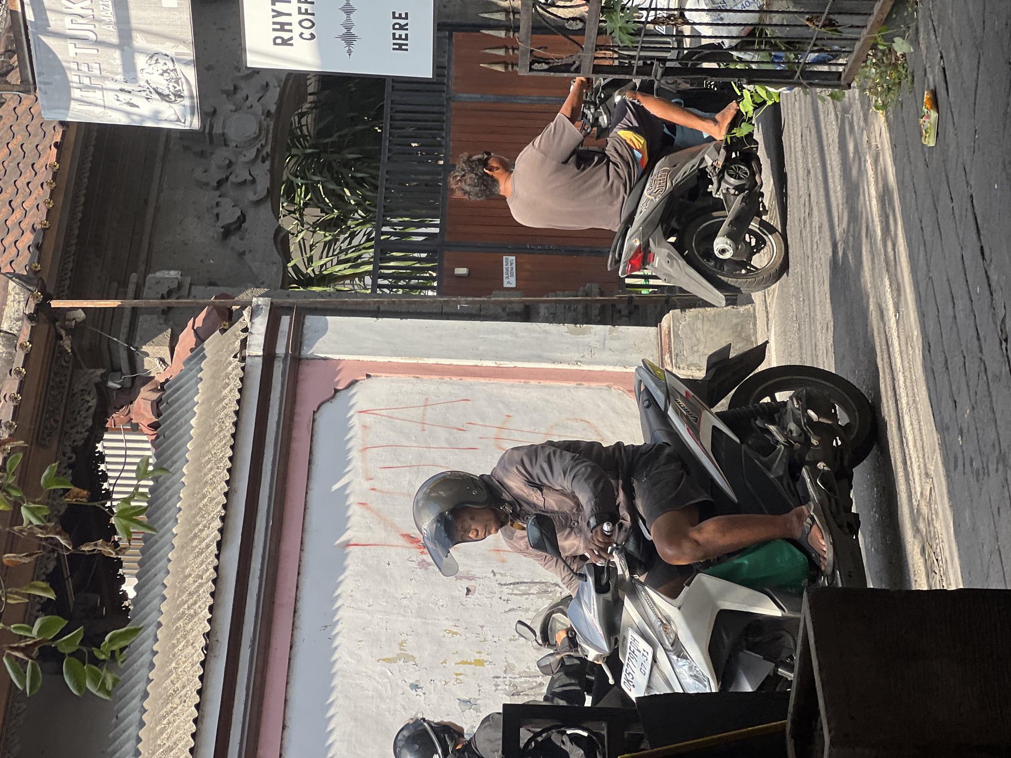 The image shows a street scene in Kuta, Indonesia, with several motorcycles and people. In the foreground, a person wearing a brown jacket and helmet is riding a white and silver scooter. The scooter has a license plate that reads "DK5779FDH". To the right, another person wearing a brown t-shirt and shorts is riding a black scooter. They are wearing sunglasses and have curly hair. In the background, there is a building with a tiled roof and a sign that says "LIFE TURK". There is also another sign with the words "RHYTHM COFFEE" and "HERE". The street is paved with dark stones and there is a small offering on the ground to the right. The scene appears to be in a commercial area with shops and buildings lining the street. The weather is sunny.